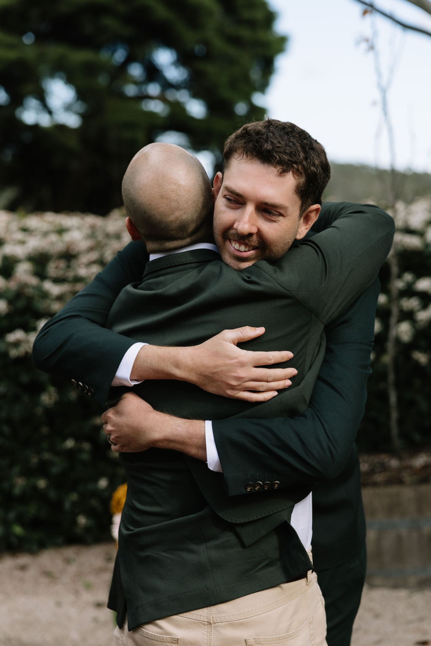 Two men in dark green suits share a warm hug at an outdoor wedding ceremony.