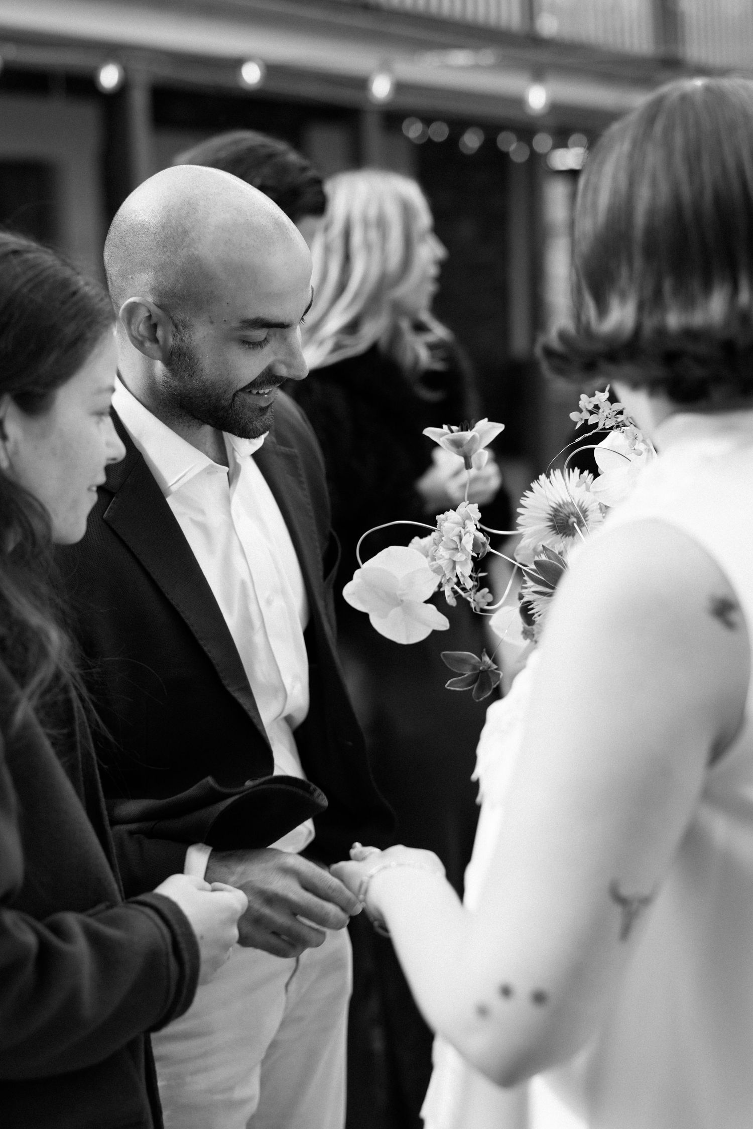 Black and white photo of a bald man in a suit smiling while exchanging rings at an outdoor wedding ceremony.