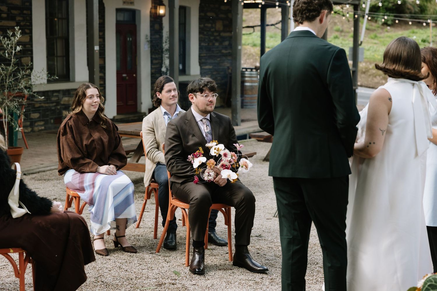 Guests seated outdoors at an intimate wedding ceremony, with the groom holding a floral bouquet in a brown suit.