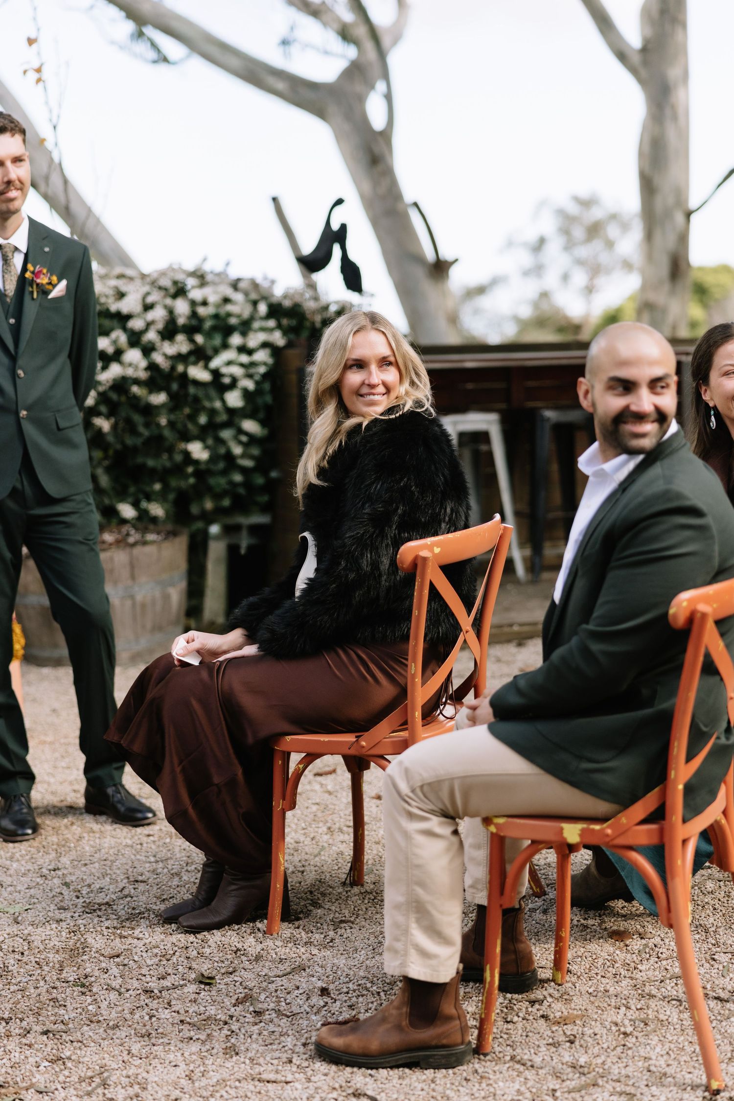 Guests smiling and seated on orange wooden chairs at an outdoor wedding ceremony surrounded by trees.