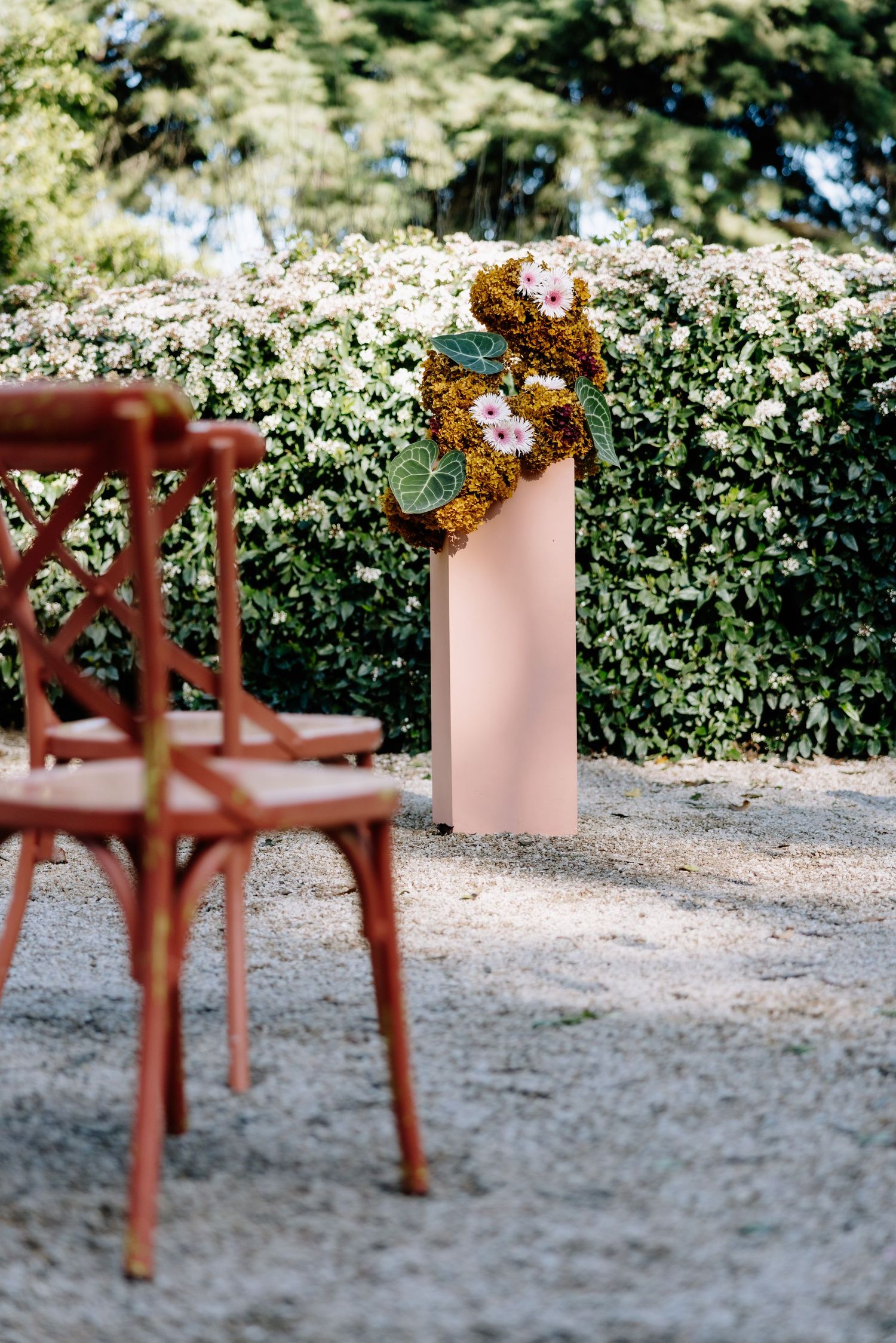 Rustic red chair beside a pink pedestal with floral arrangement against a lush green hedge at an outdoor wedding.
