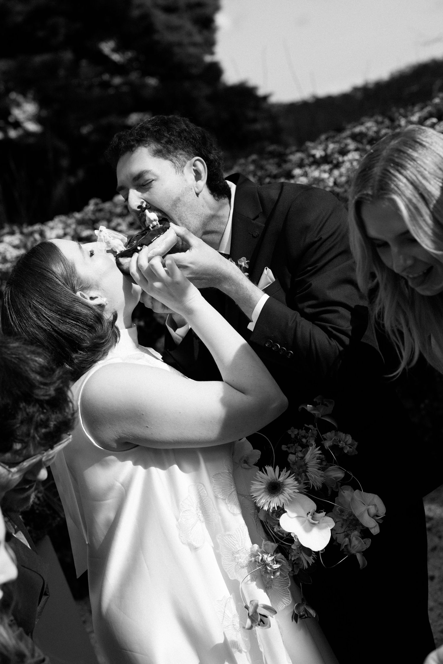 Black and white photo of bride and groom sharing wedding cake, with guest nearby amid floral arrangements.