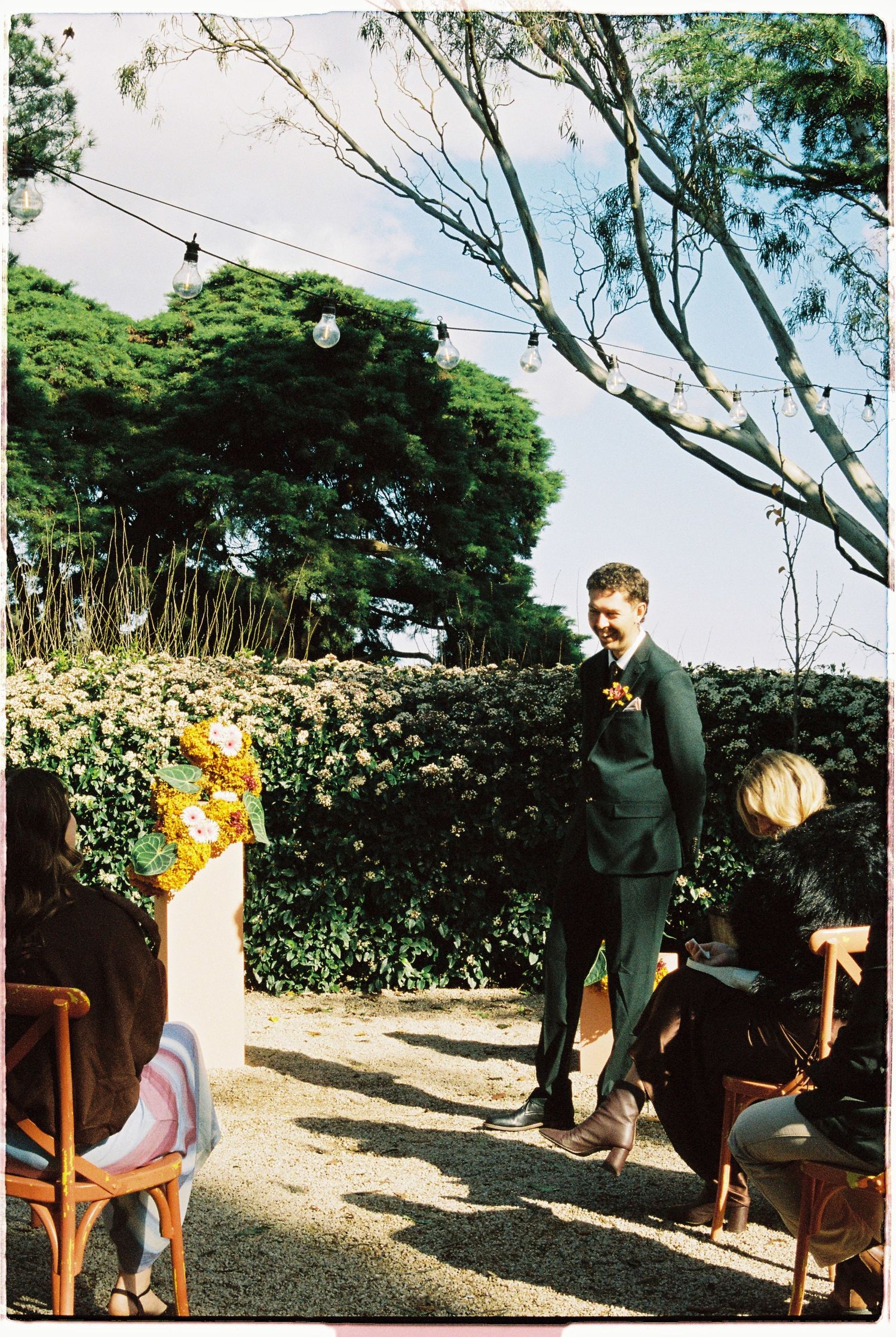 Groom in green suit stands at outdoor wedding ceremony surrounded by floral arrangements and string lights.