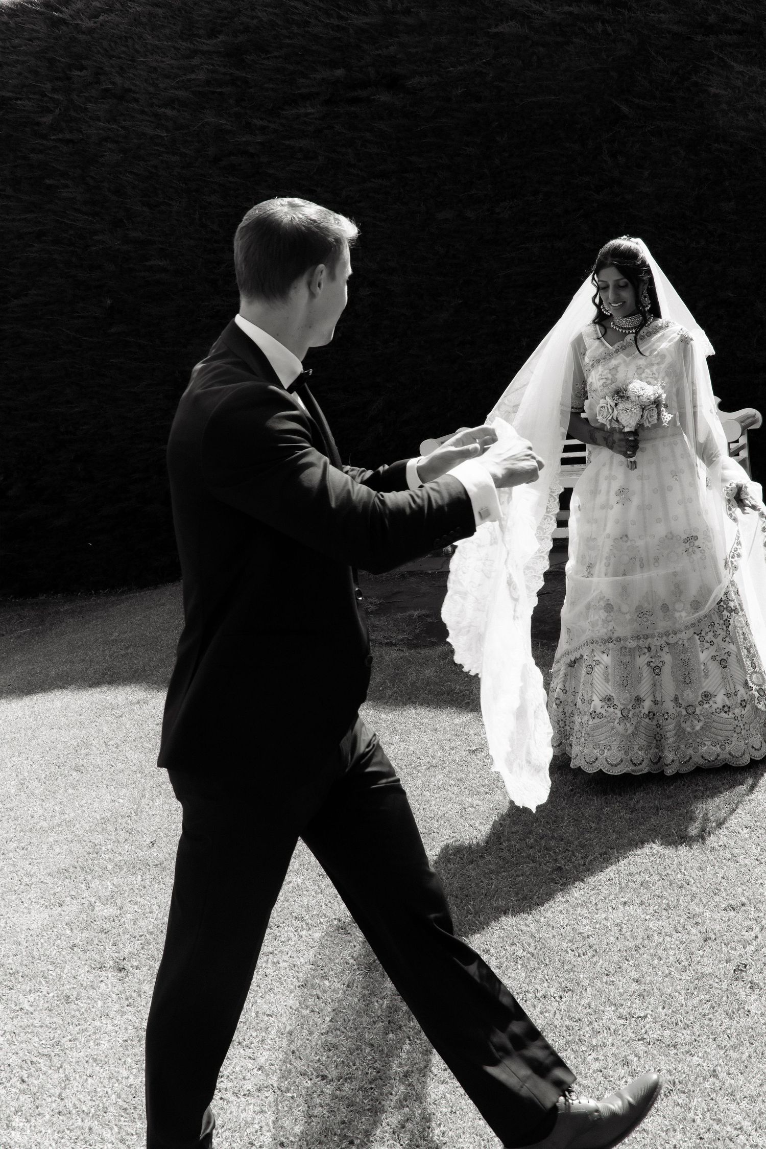 Black and white photo of groom reaching out to bride in lace wedding dress and veil outdoors.