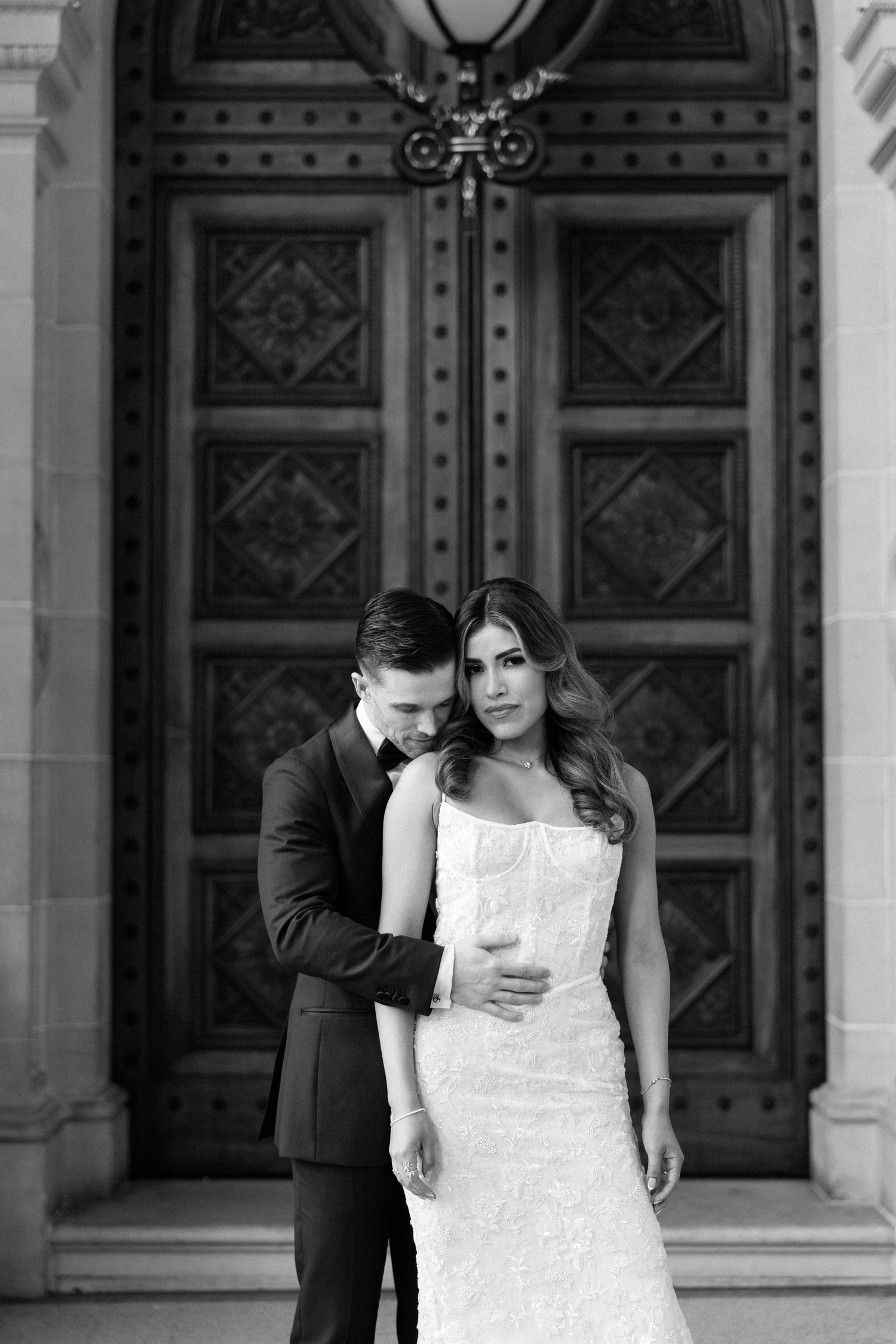 Black and white portrait of groom embracing bride in lace dress before ornate wooden doors.