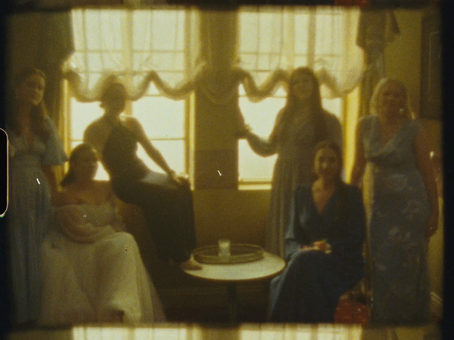Vintage photo of six women in elegant gowns gathered in a warmly lit room with draped windows.