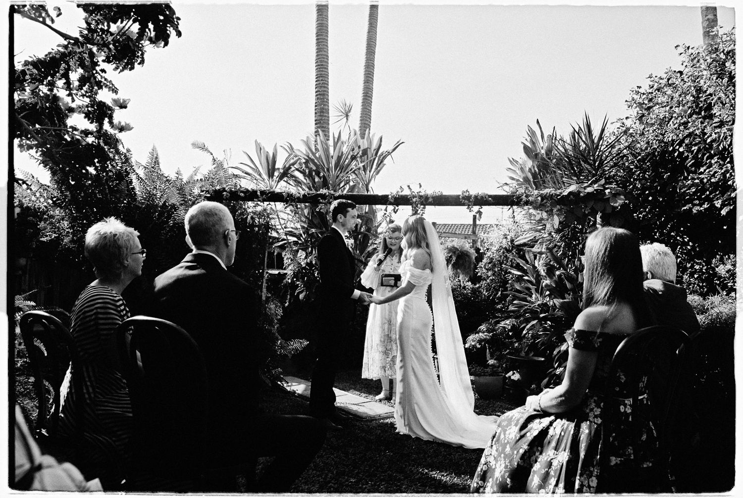 Outdoor garden wedding ceremony with bride and groom at floral arch surrounded by seated guests.
