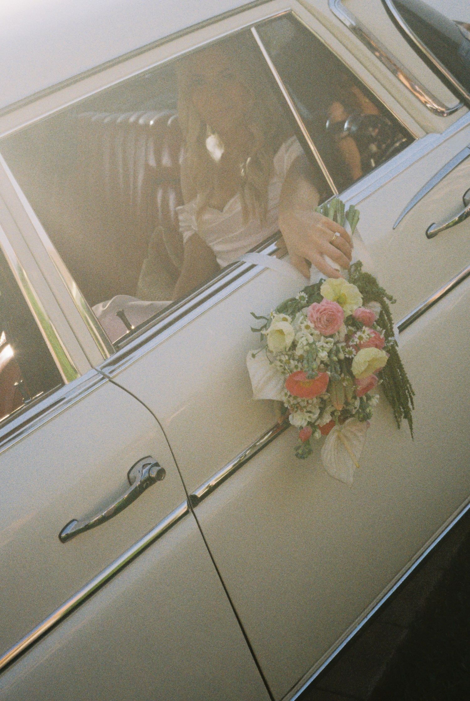 Bride holding colorful bouquet through vintage car window on wedding day.