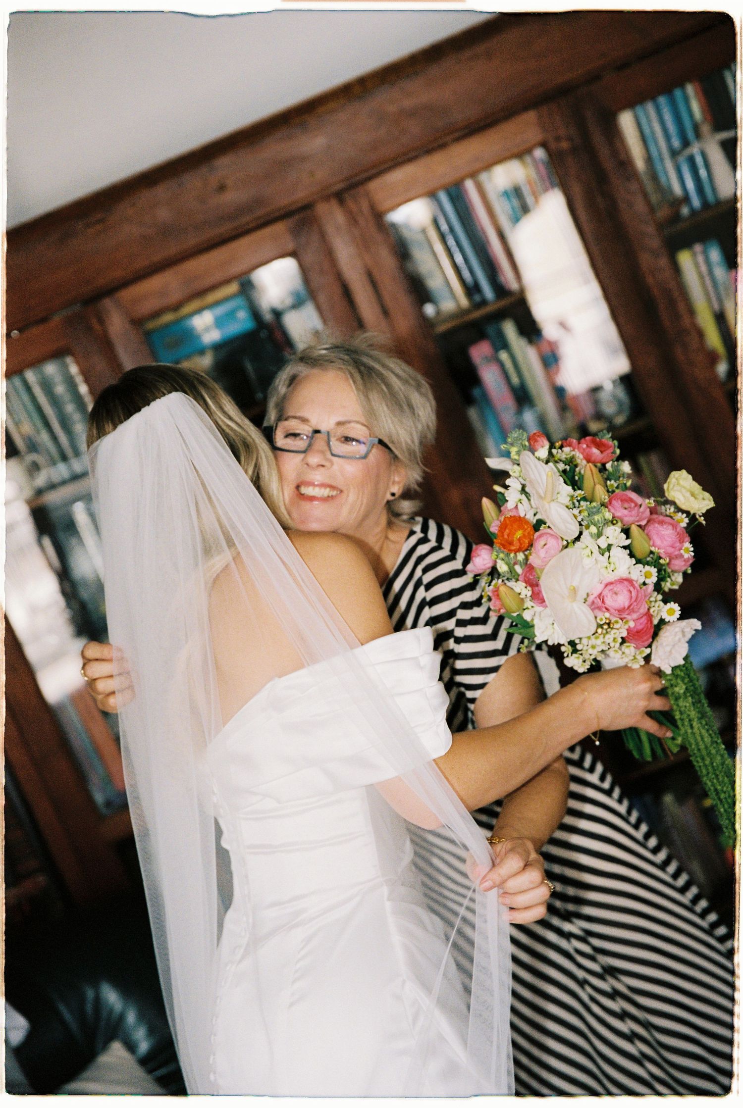 Smiling woman in glasses hugs bride in white gown and veil while holding colorful floral bouquet indoors.