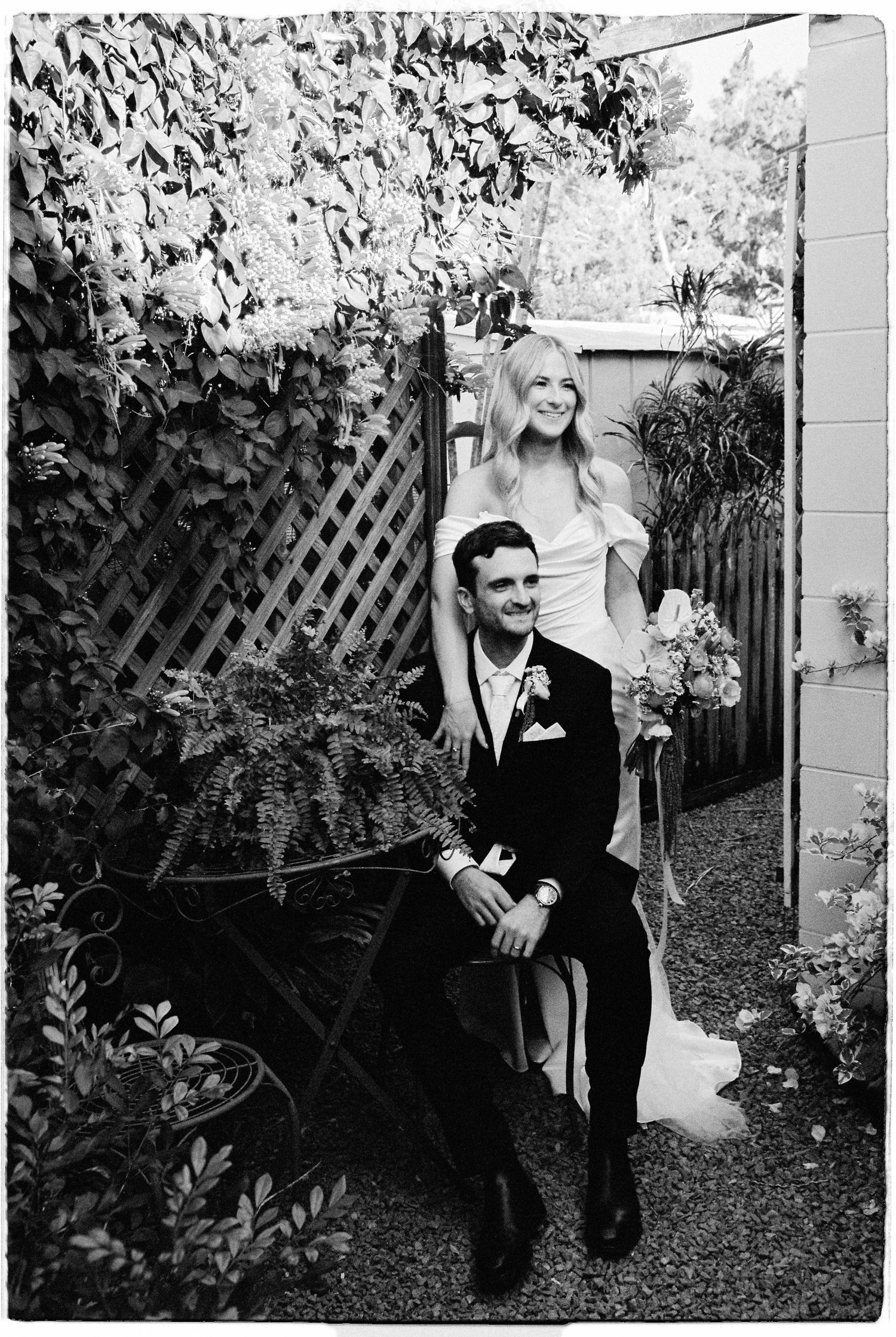 Black and white photo of bride standing behind seated groom in lush garden setting.