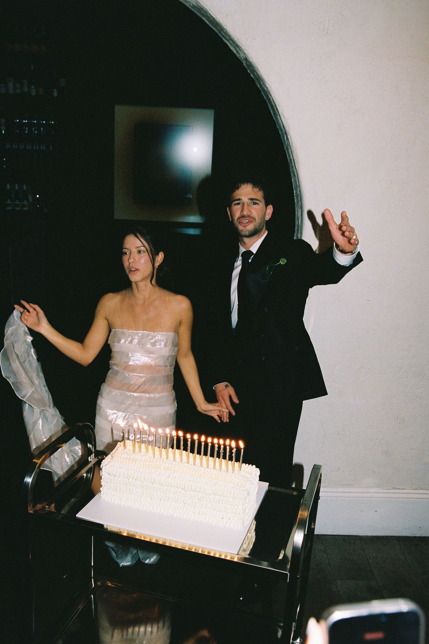 Wedding couple standing behind white cake with candles, groom raising hand in celebration at reception.