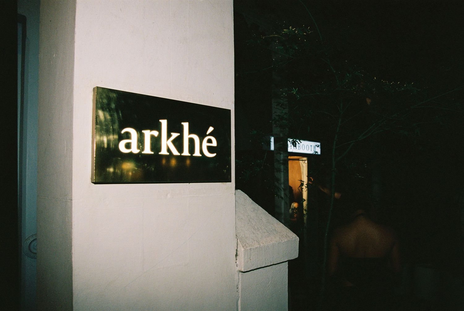Arkhe restaurant entrance sign mounted on a white wall in a dimly lit outdoor setting at night.