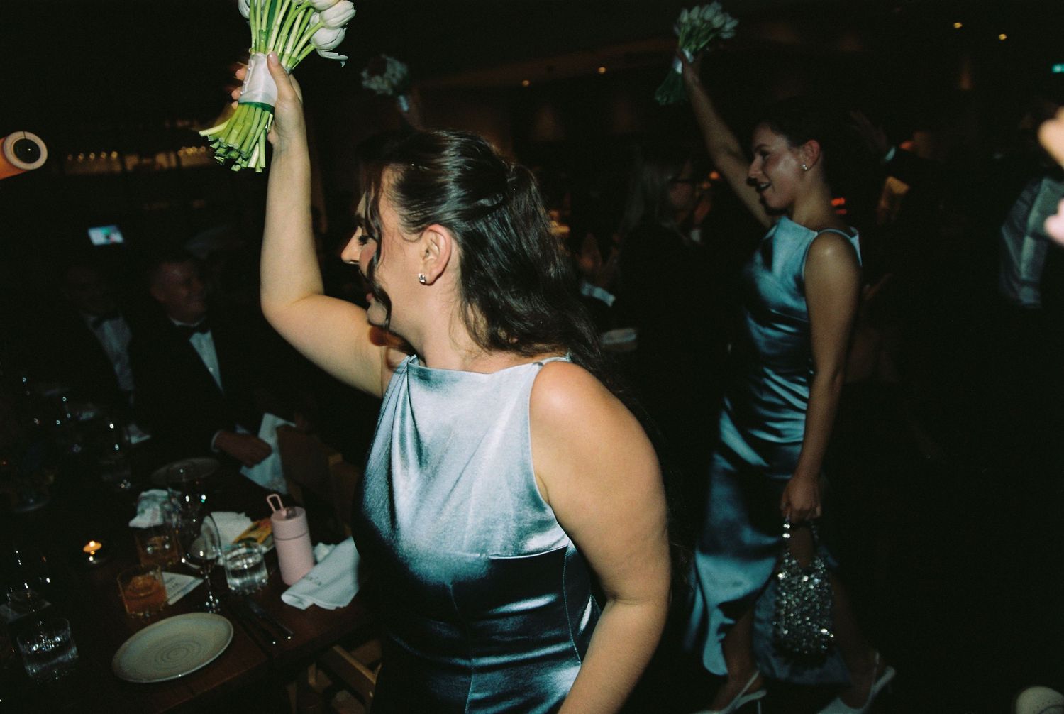 Wedding guest in blue satin dress triumphantly holding a white bridal bouquet.