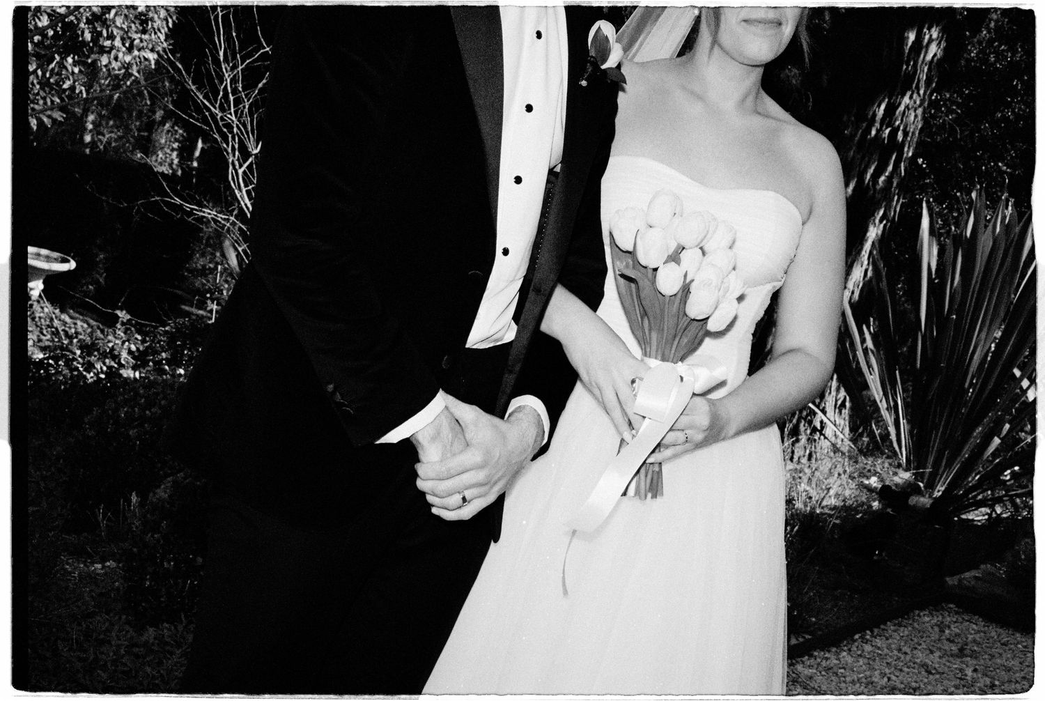 Black and white close-up of bride holding tulip bouquet while holding hands with groom outdoors.