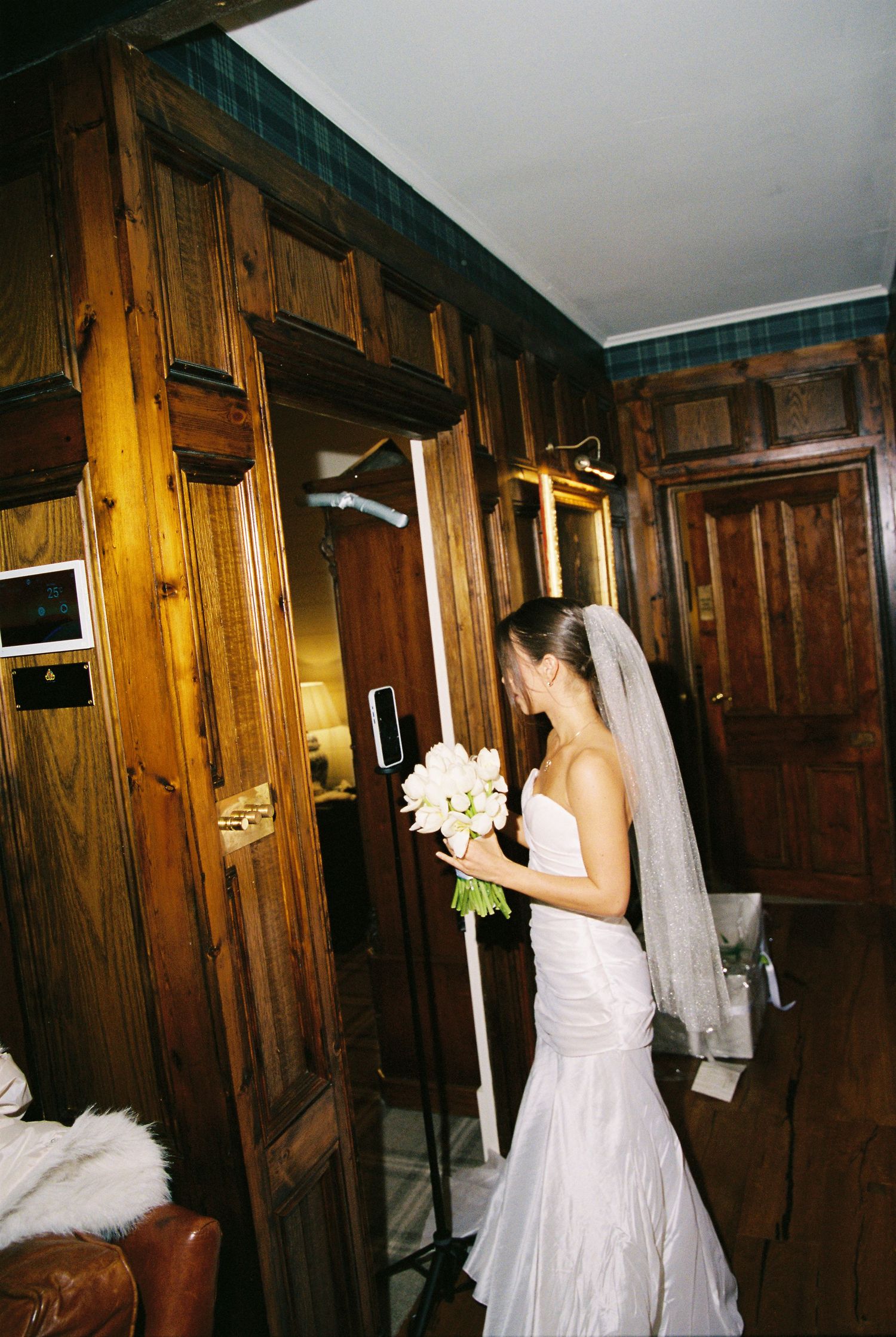 Bride in white wedding gown holding bouquet standing in a dark wood-paneled room.