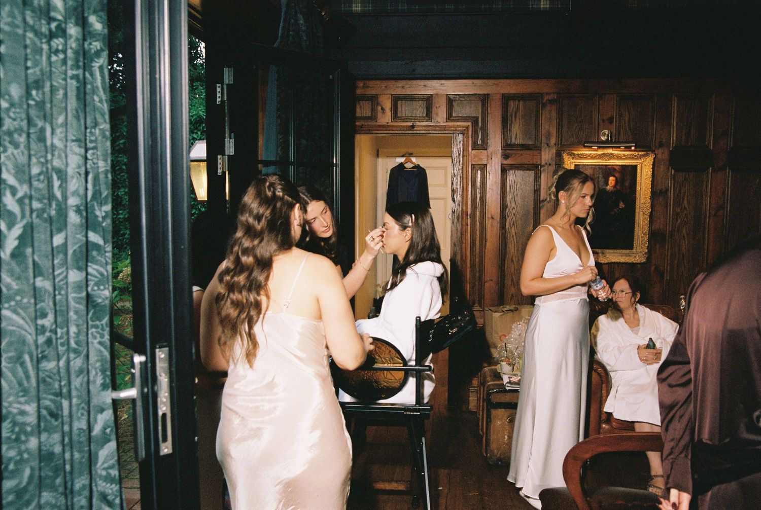 Bridesmaids in satin dresses getting ready together in wood-paneled dressing room.