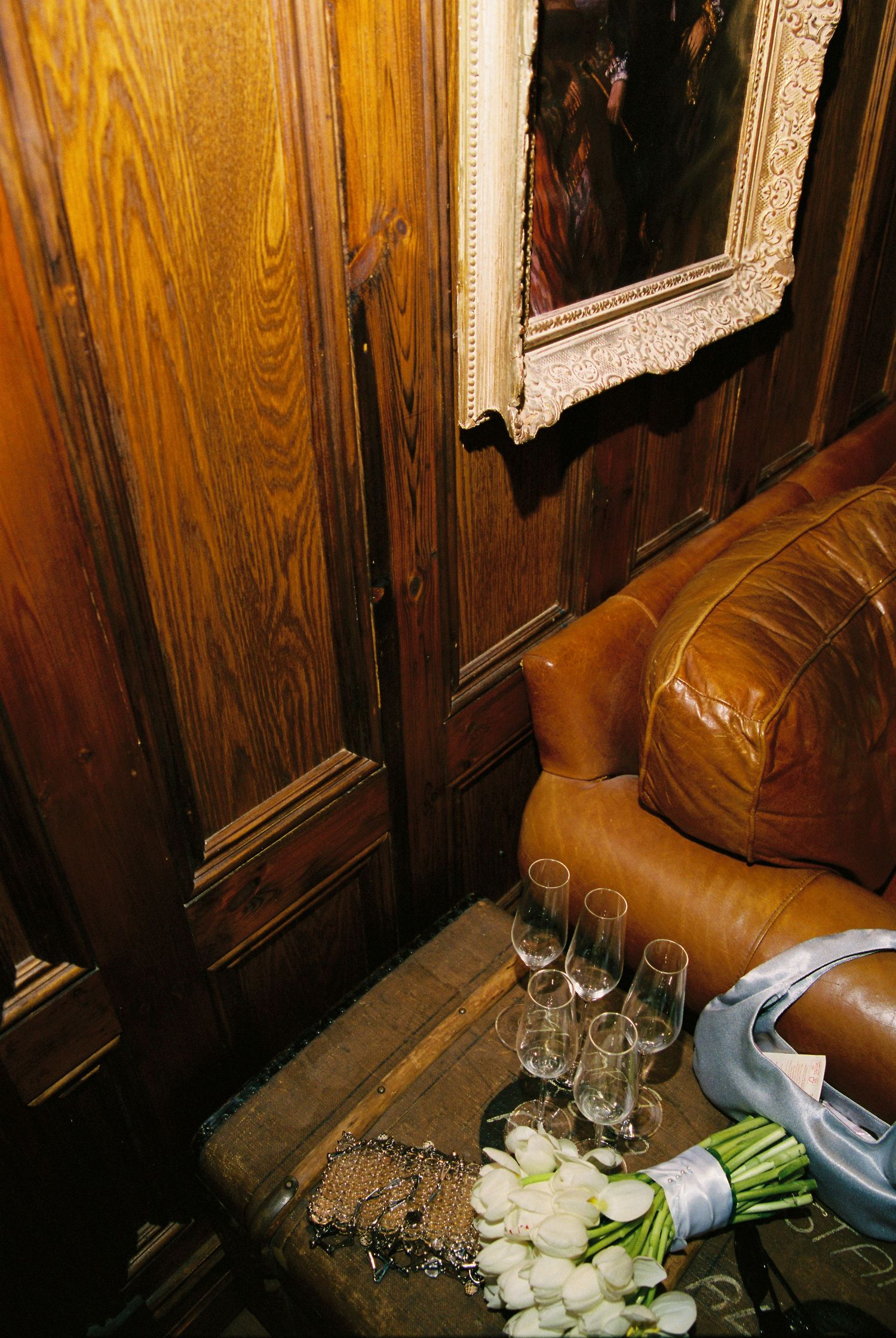Champagne glasses and white tulip bouquet on vintage trunk beside leather sofa in wood-paneled room.