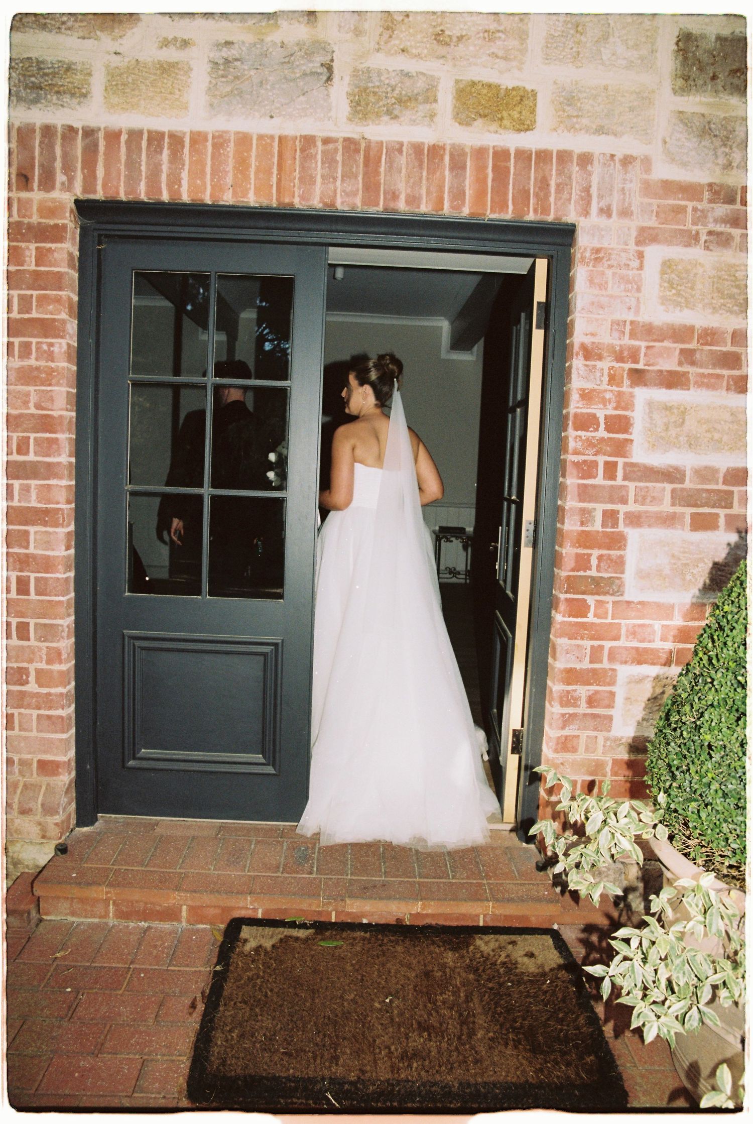 Bride in strapless white tulle gown and veil stands in doorway of rustic brick building at wedding.