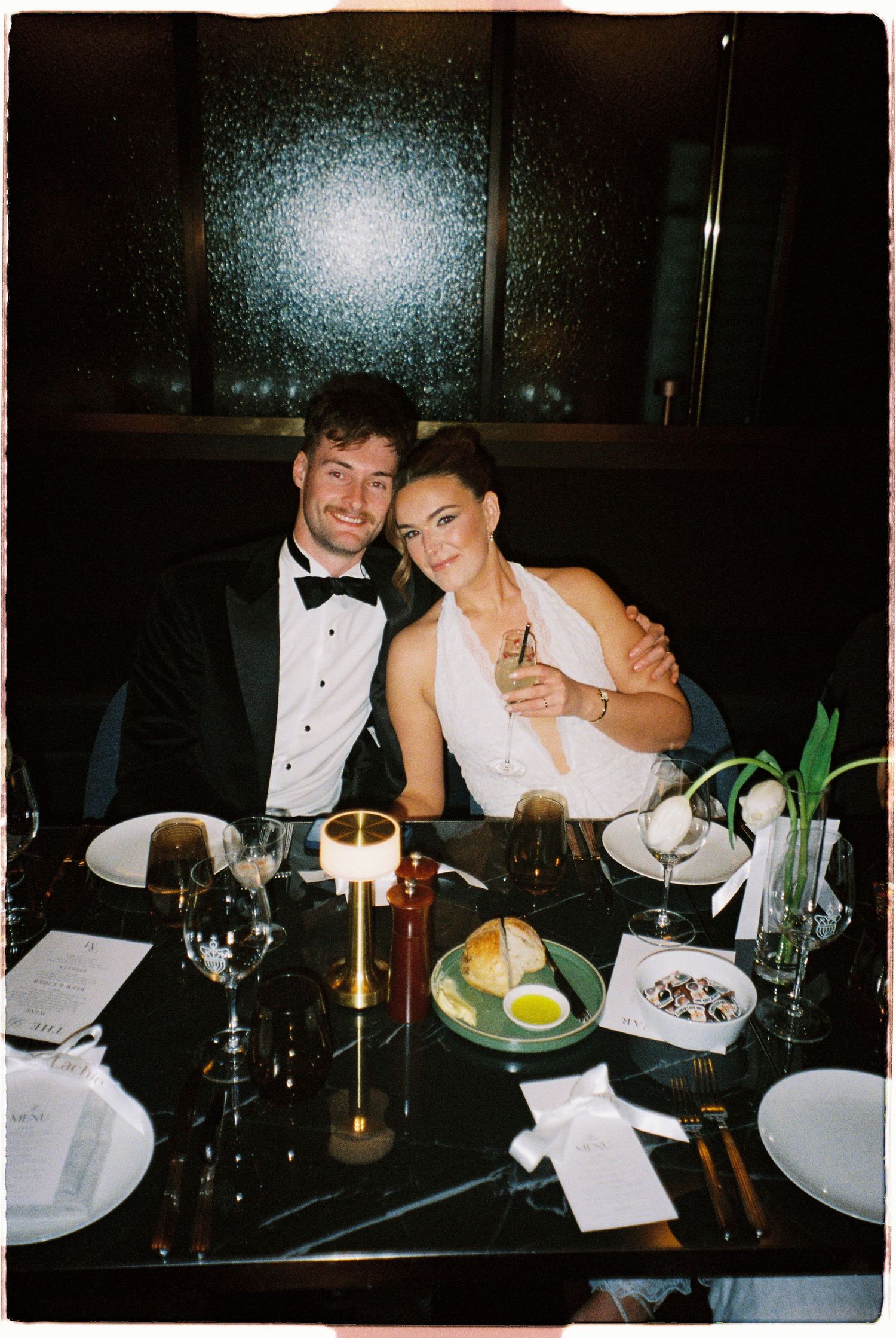 Newlywed couple smiles and embraces at candlelit restaurant dinner table during wedding reception celebration.