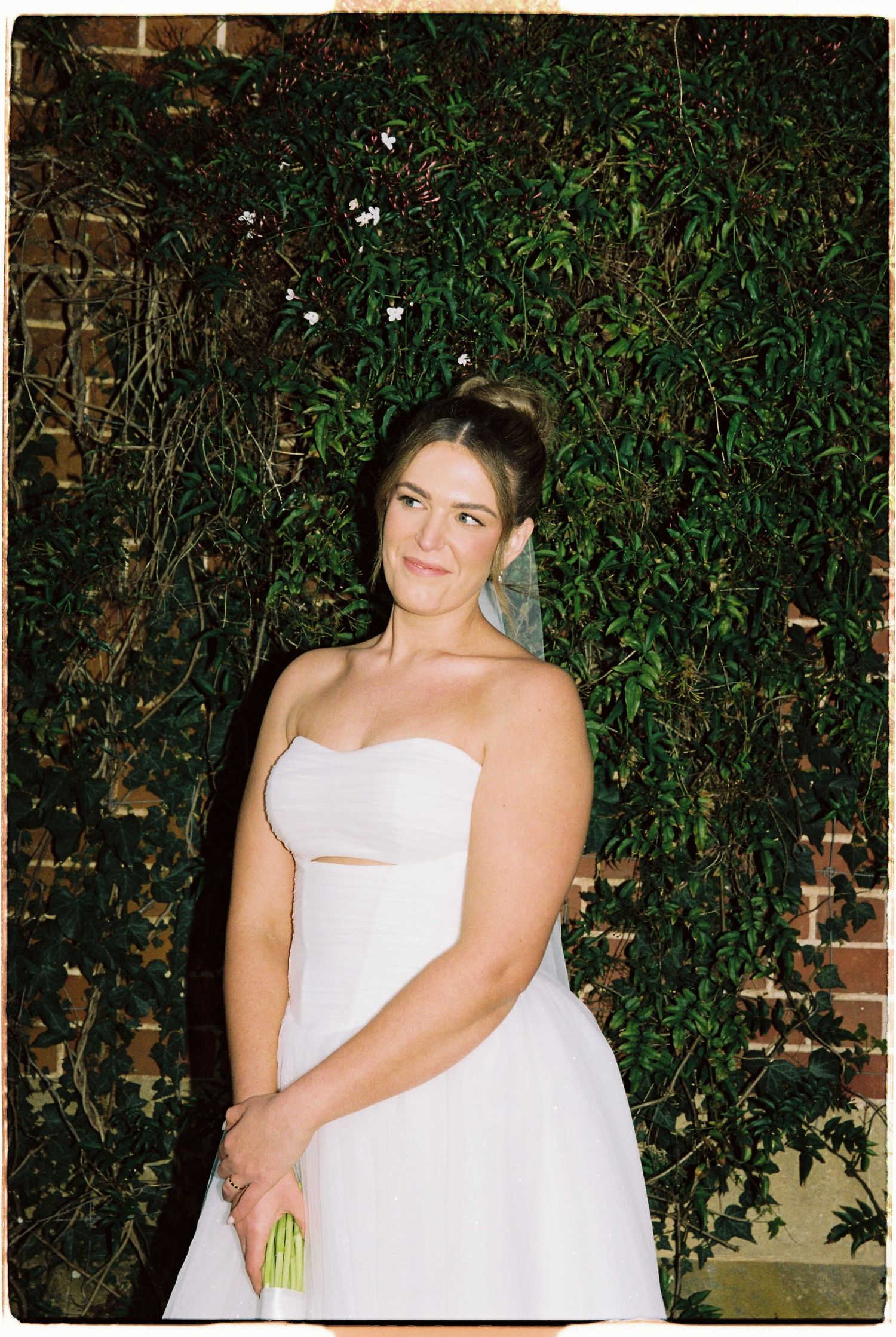 Bride in strapless white wedding gown holding bouquet, posing in front of lush green ivy wall at night.