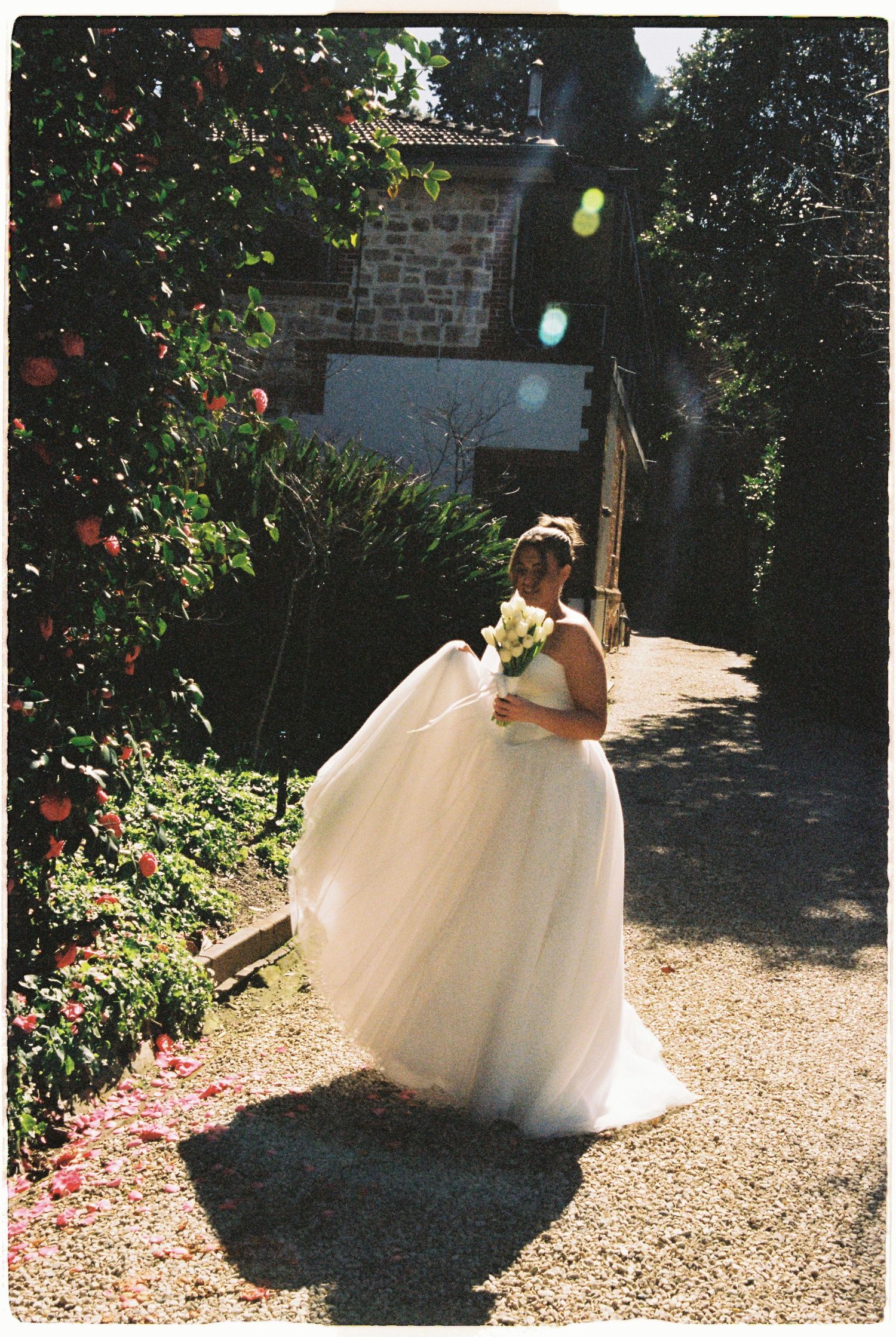 Bride in a flowing white tulle wedding dress holding a bouquet on a sunlit garden path.