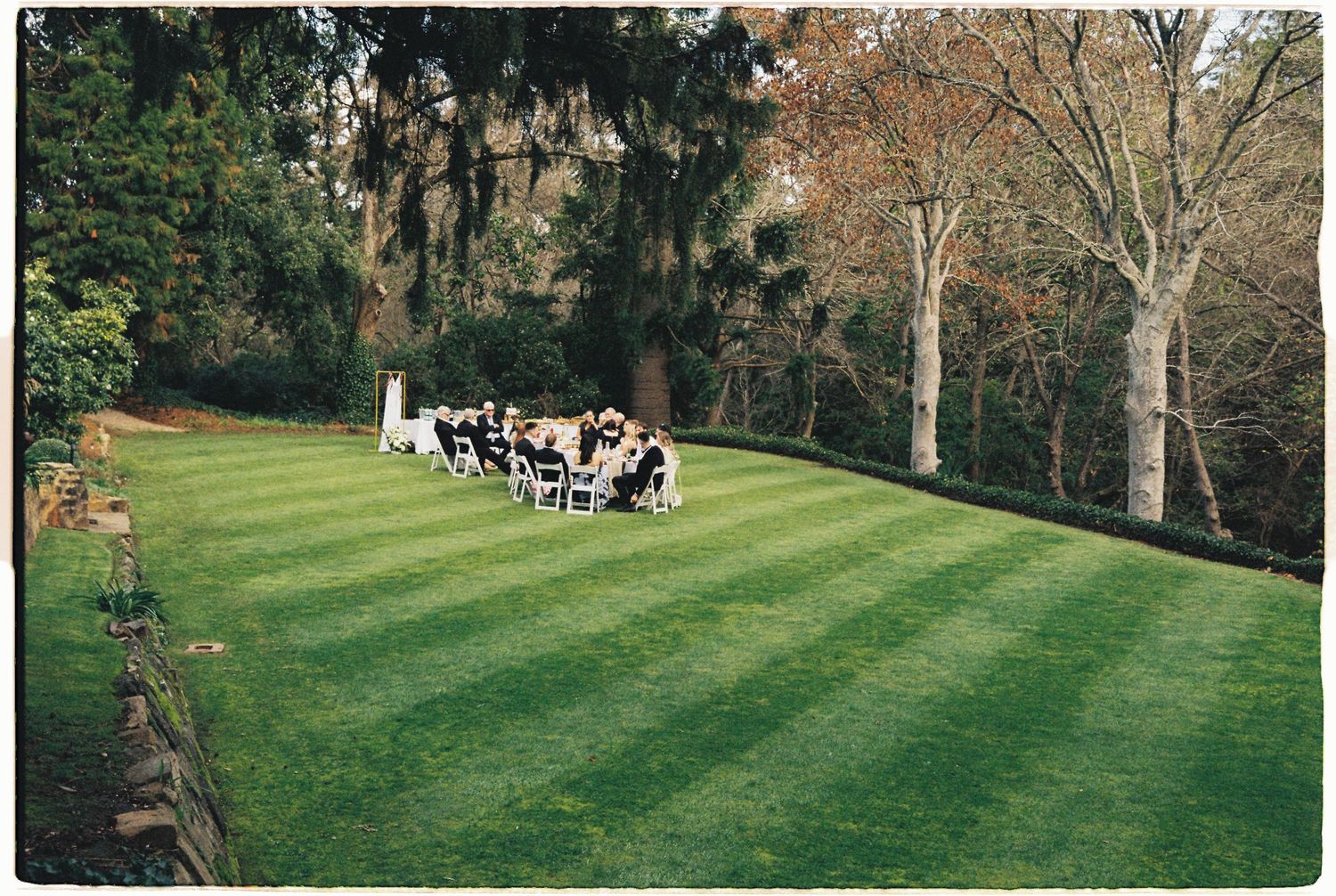 Outdoor wedding ceremony on a lush green lawn surrounded by trees and gardens.