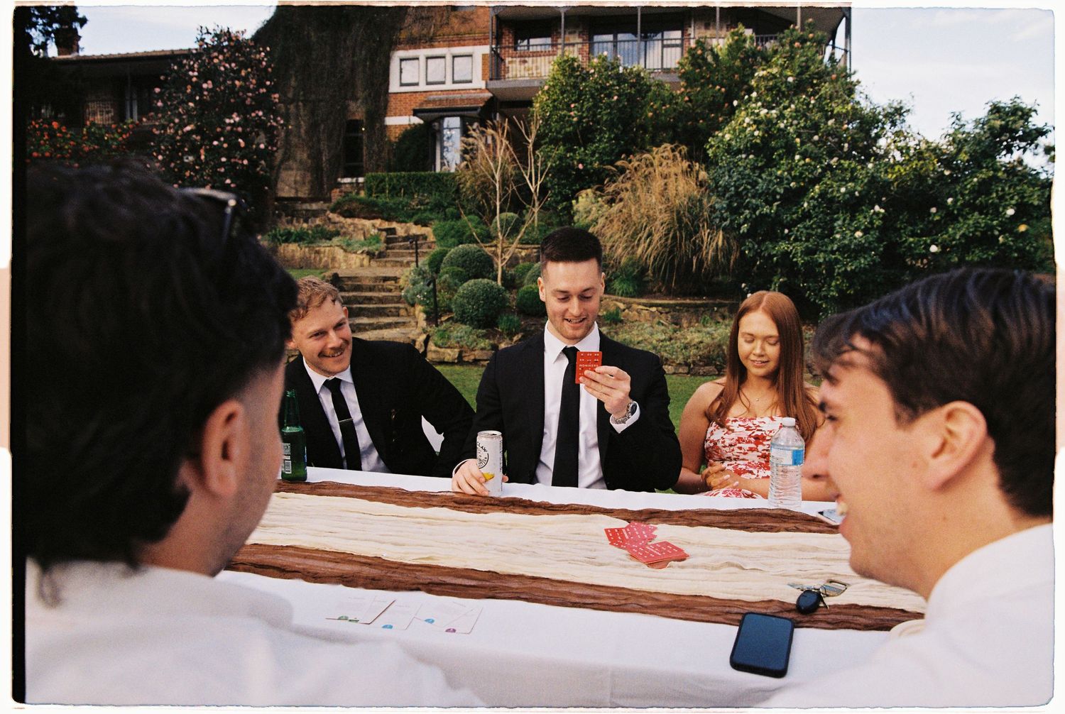 Wedding guests laughing and socializing at rustic wooden table in garden setting.