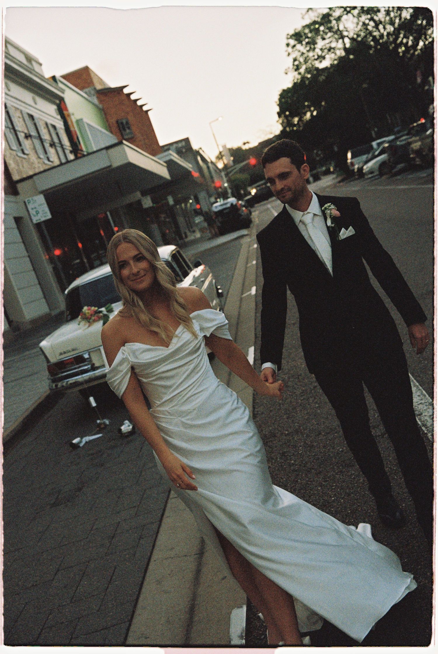 Bride in white off-shoulder gown and groom in black tuxedo hold hands walking on a city street.