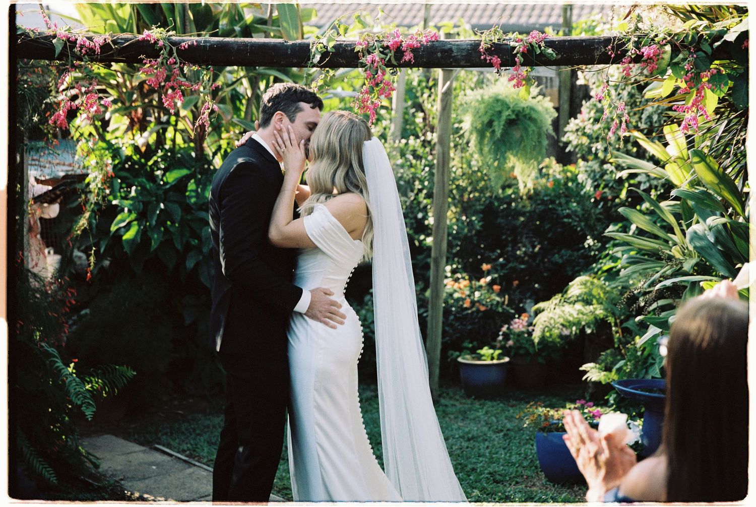 Bride and groom share first kiss under floral arch in lush tropical garden wedding ceremony.