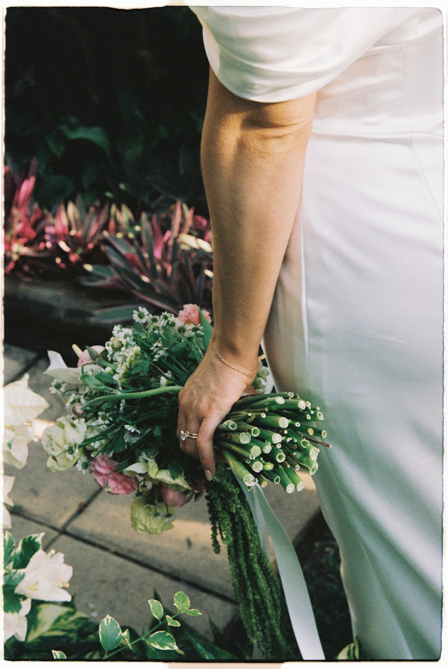 Bride holding elegant green botanical wedding bouquet with trailing ribbon against white gown outdoors.