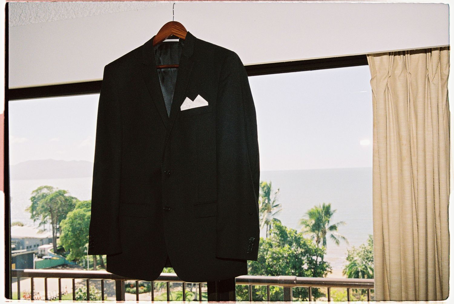 Black tuxedo jacket with white pocket square hanging on wooden hanger near balcony with ocean and palm tree view.