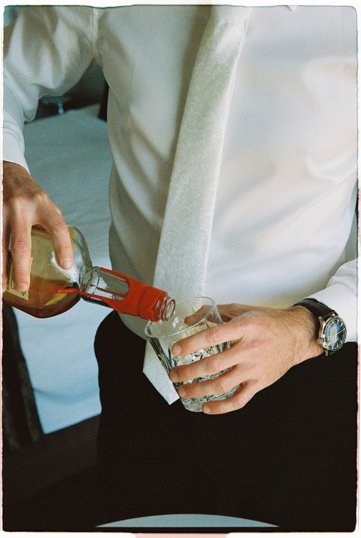 Man in white shirt and tie pouring whiskey from a bottle into a glass while wearing a watch.