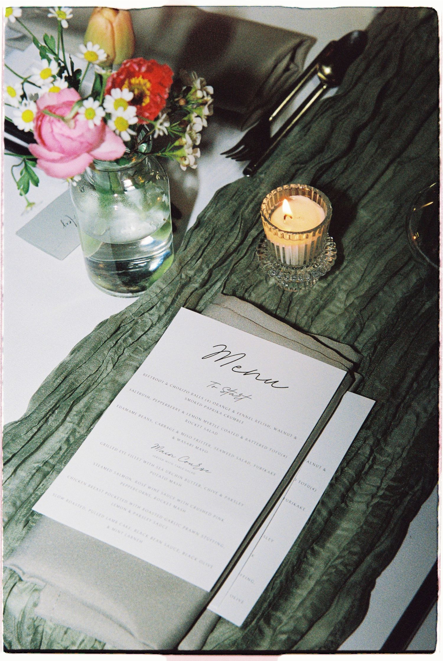Elegant wedding menu card on green linen with candle, floral arrangement, and place setting.
