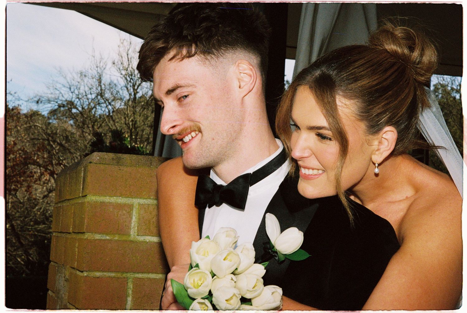 A smiling couple dressed in formal attire poses closely together outdoors, the man holding white roses.