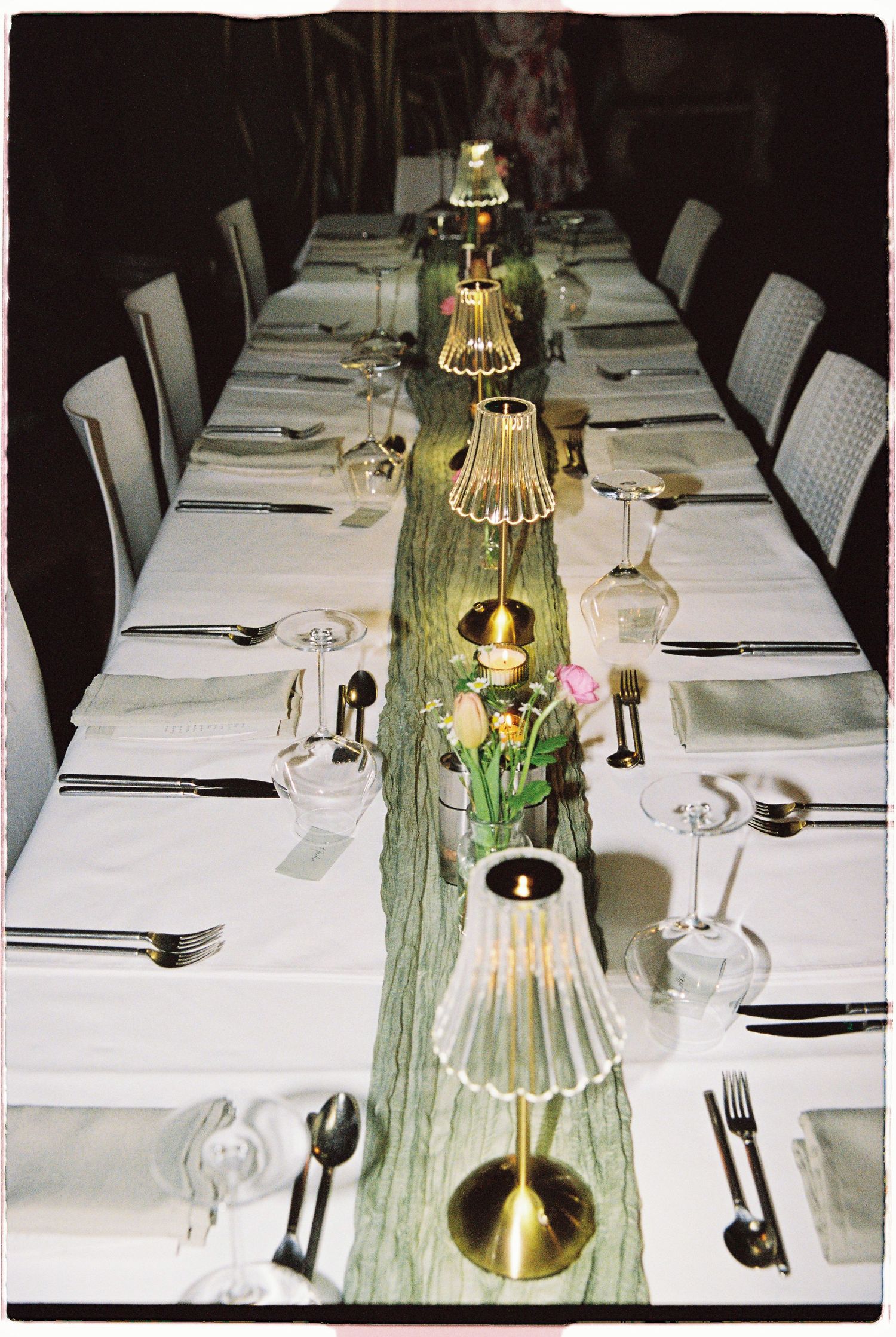 Elegant long dining table set with white linens, gold candle holders, glassware, and a green runner for a formal dinner.