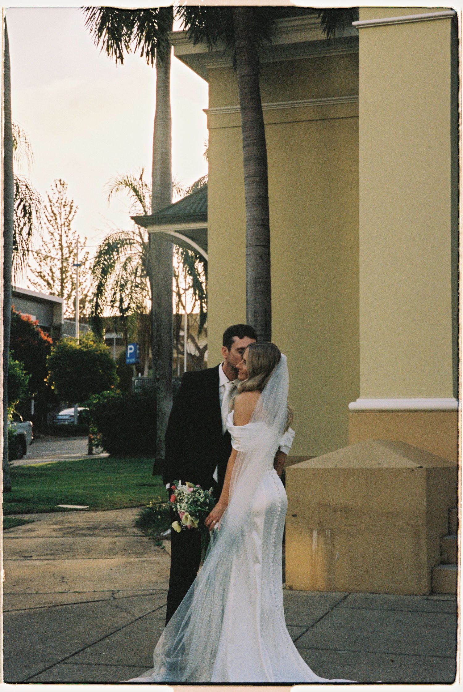 Bride and groom share a romantic kiss outside a yellow building surrounded by palm trees on their wedding day.