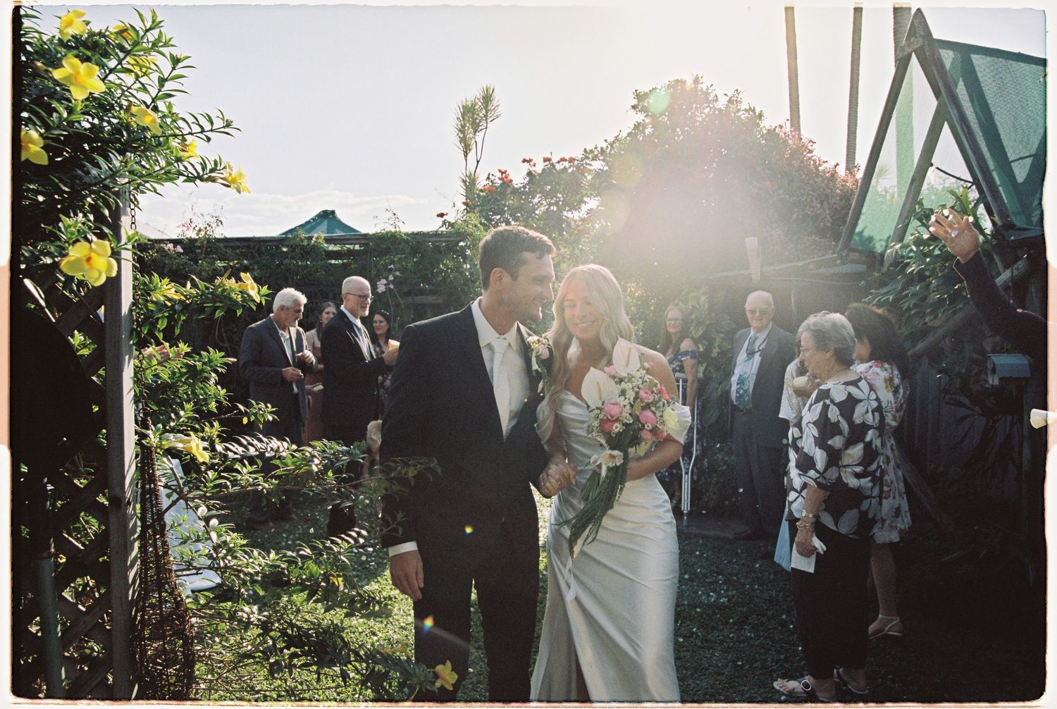 Newlyweds share a romantic kiss bathed in golden sunlight surrounded by wedding guests in garden setting.