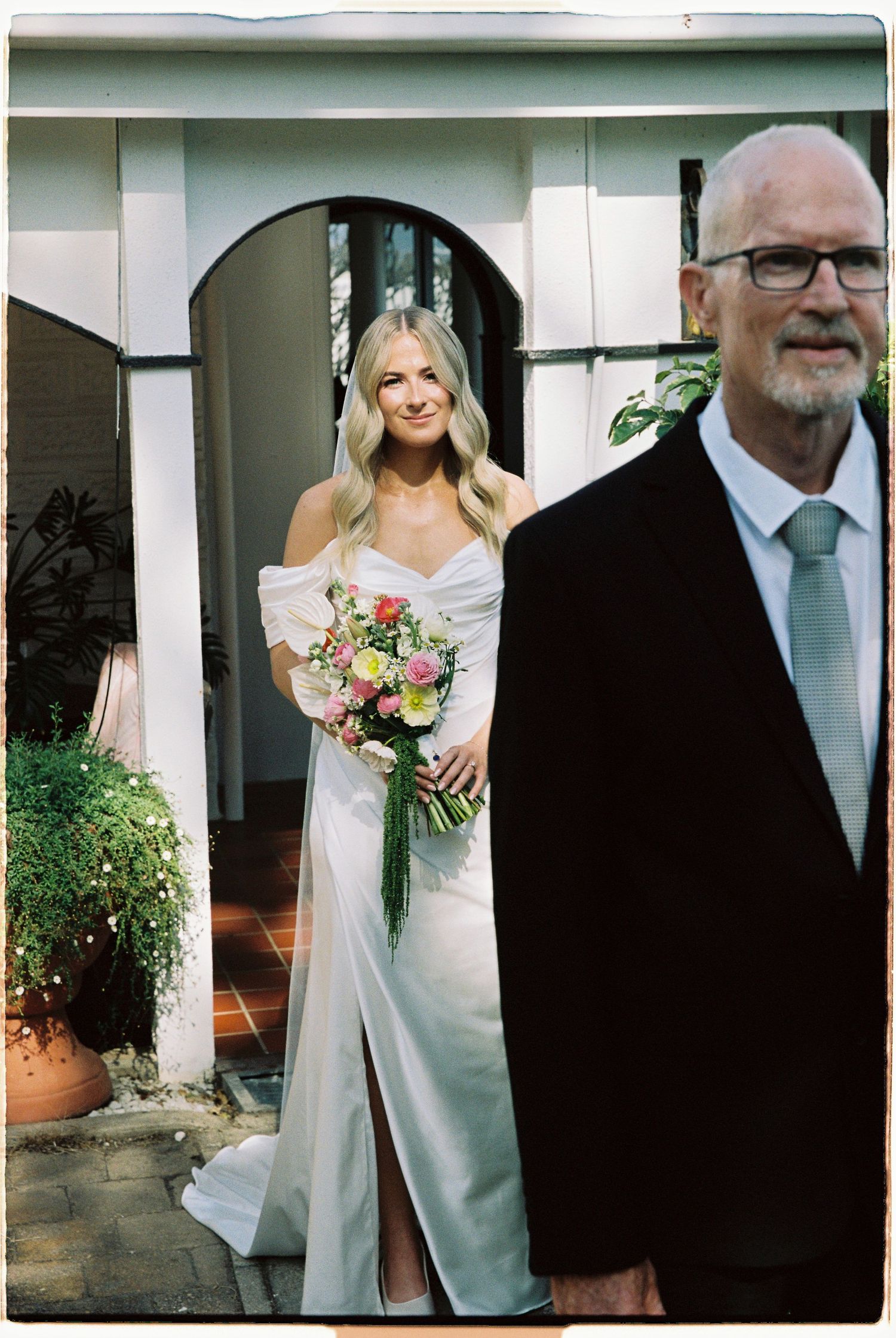 Bride in white off-shoulder gown holding colorful bouquet stands in arched doorway beside older man in black suit.