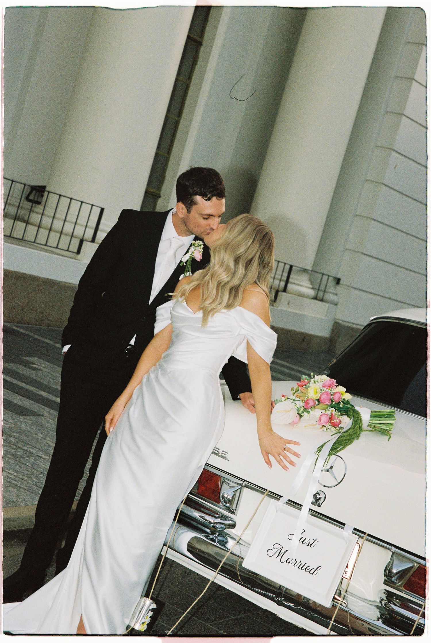 Newlywed couple sharing a kiss beside a white Just Married car outside a church after their wedding ceremony.