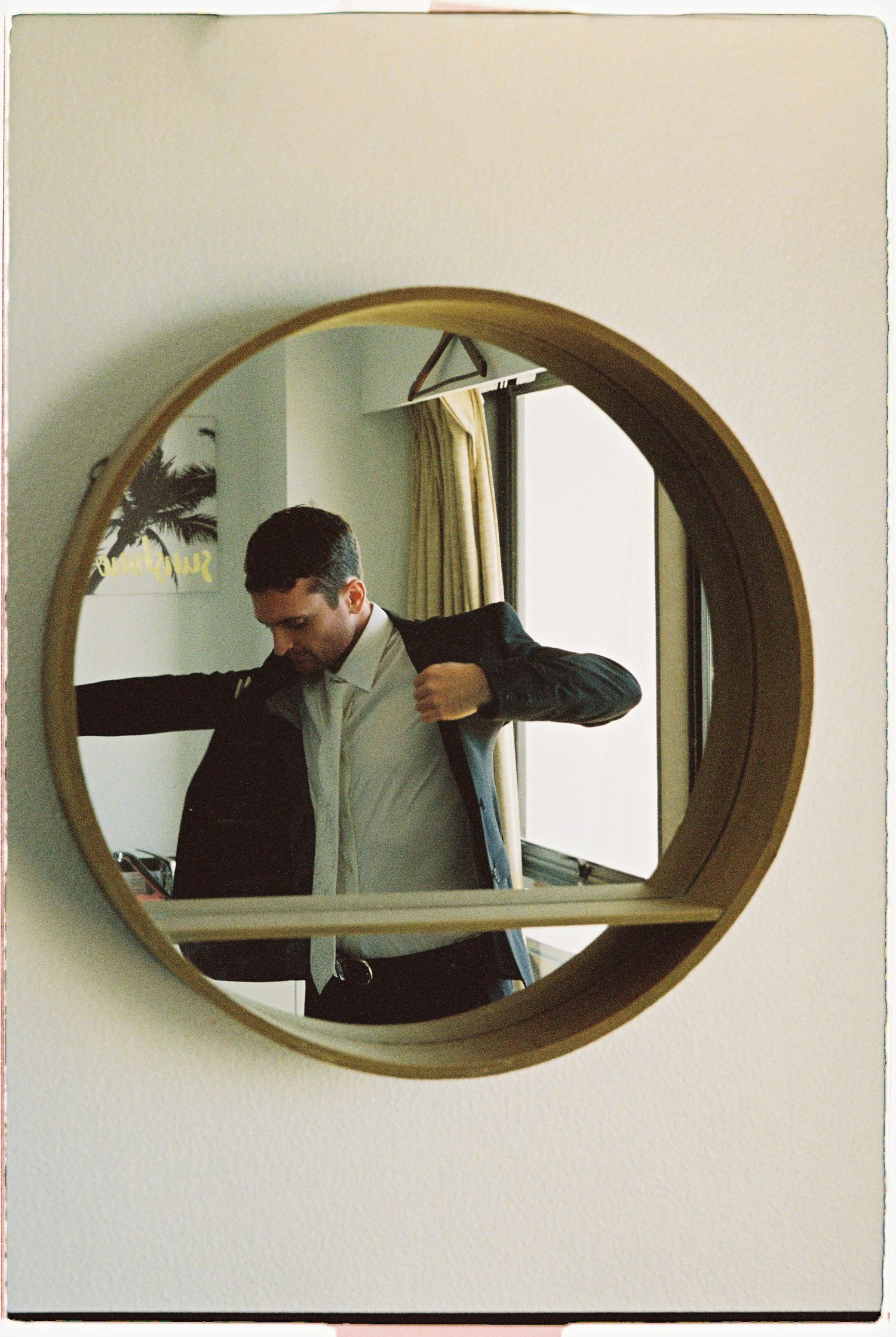 Groom putting on suit jacket reflected in round gold-framed wall mirror in hotel room.