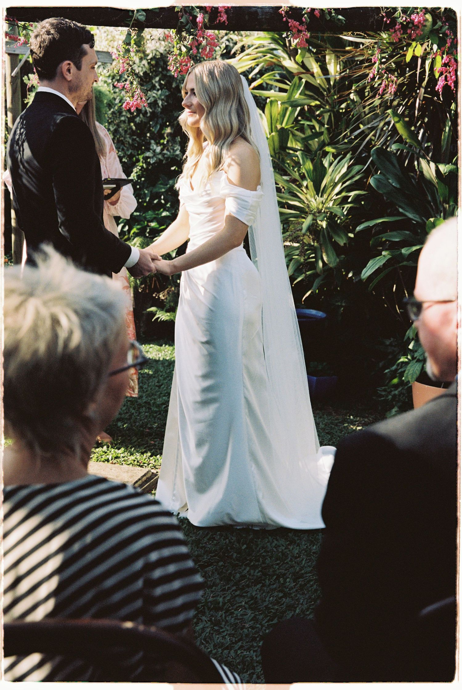 Bride in white gown holding hands with groom at outdoor garden ceremony.