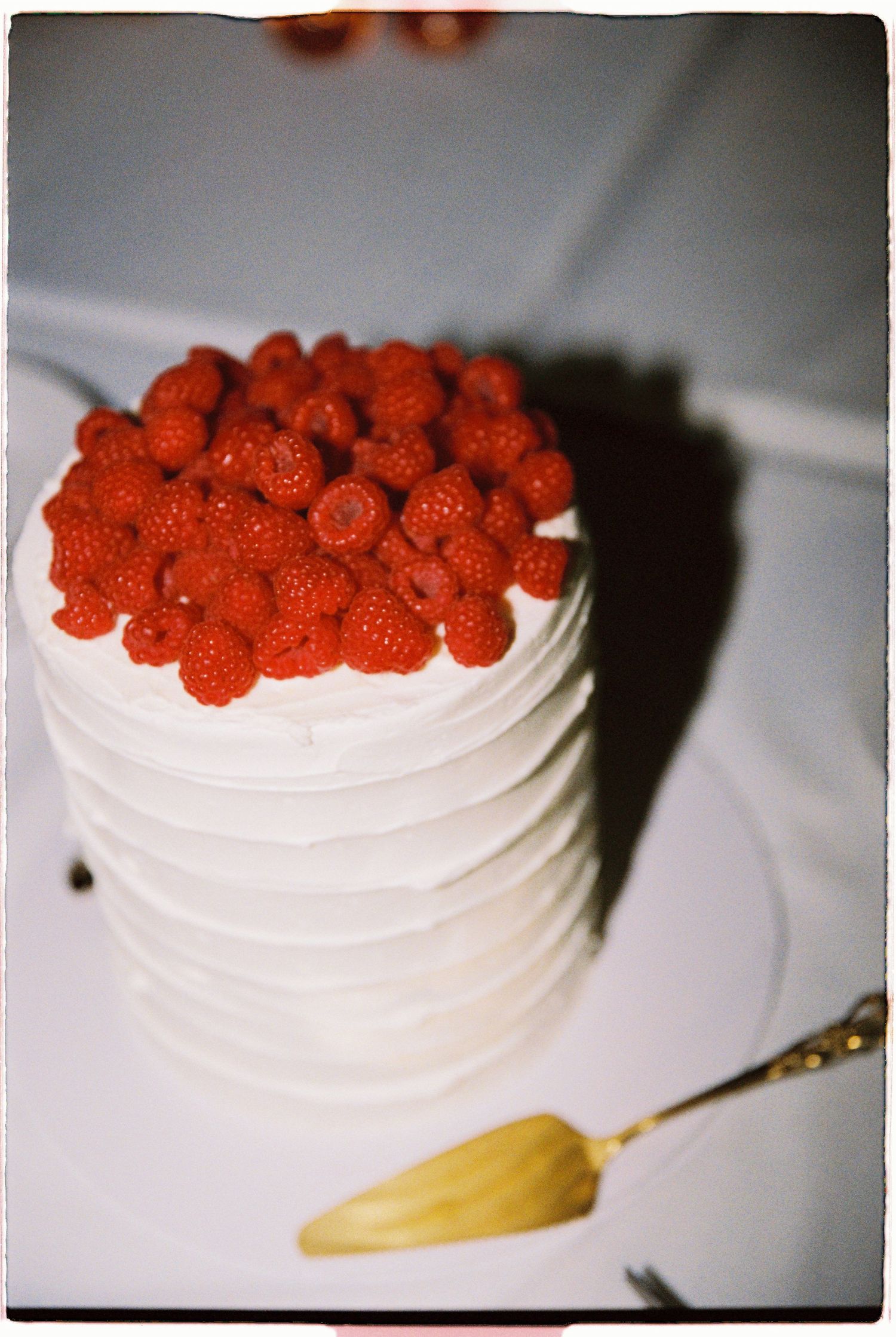White layered wedding cake generously topped with fresh red raspberries on white cake stand with gold server.