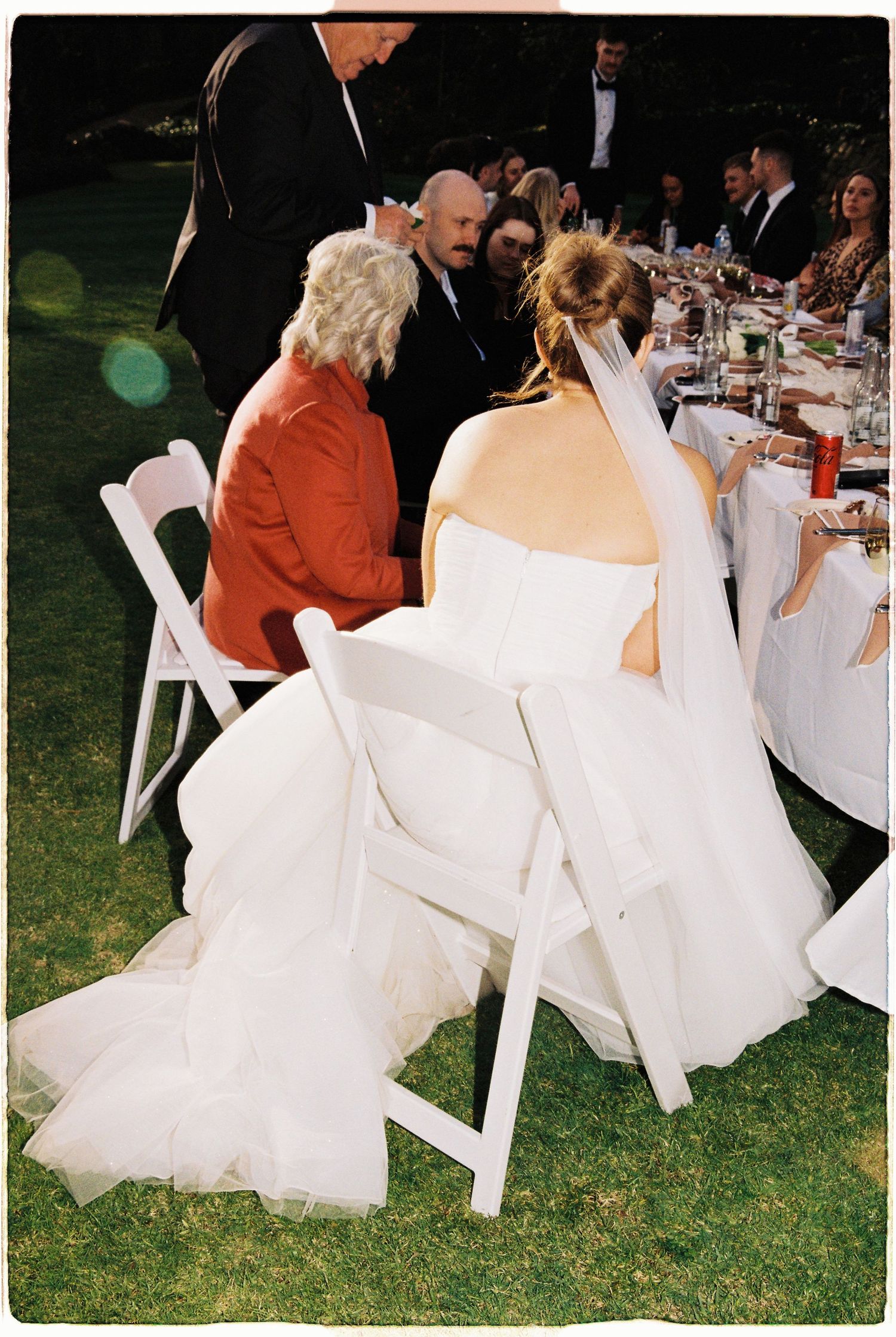 Bride in white gown seated at outdoor wedding reception table with guests nearby.