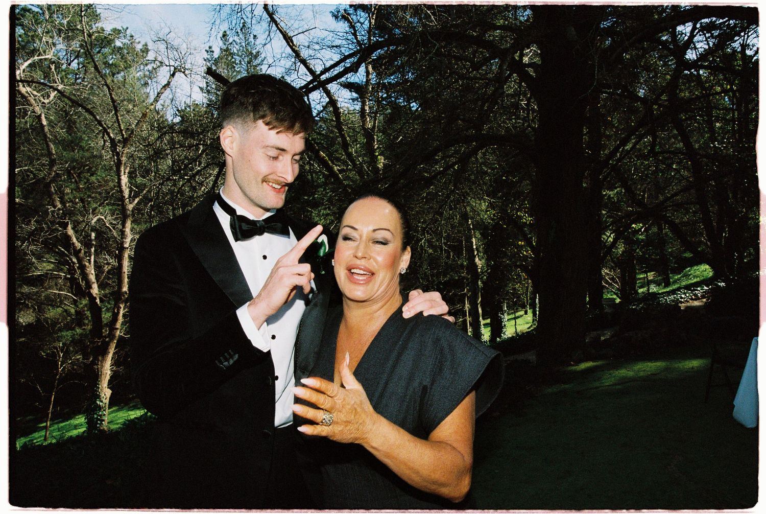 Groom in black tuxedo poses playfully with smiling woman in black dress at outdoor wedding reception.