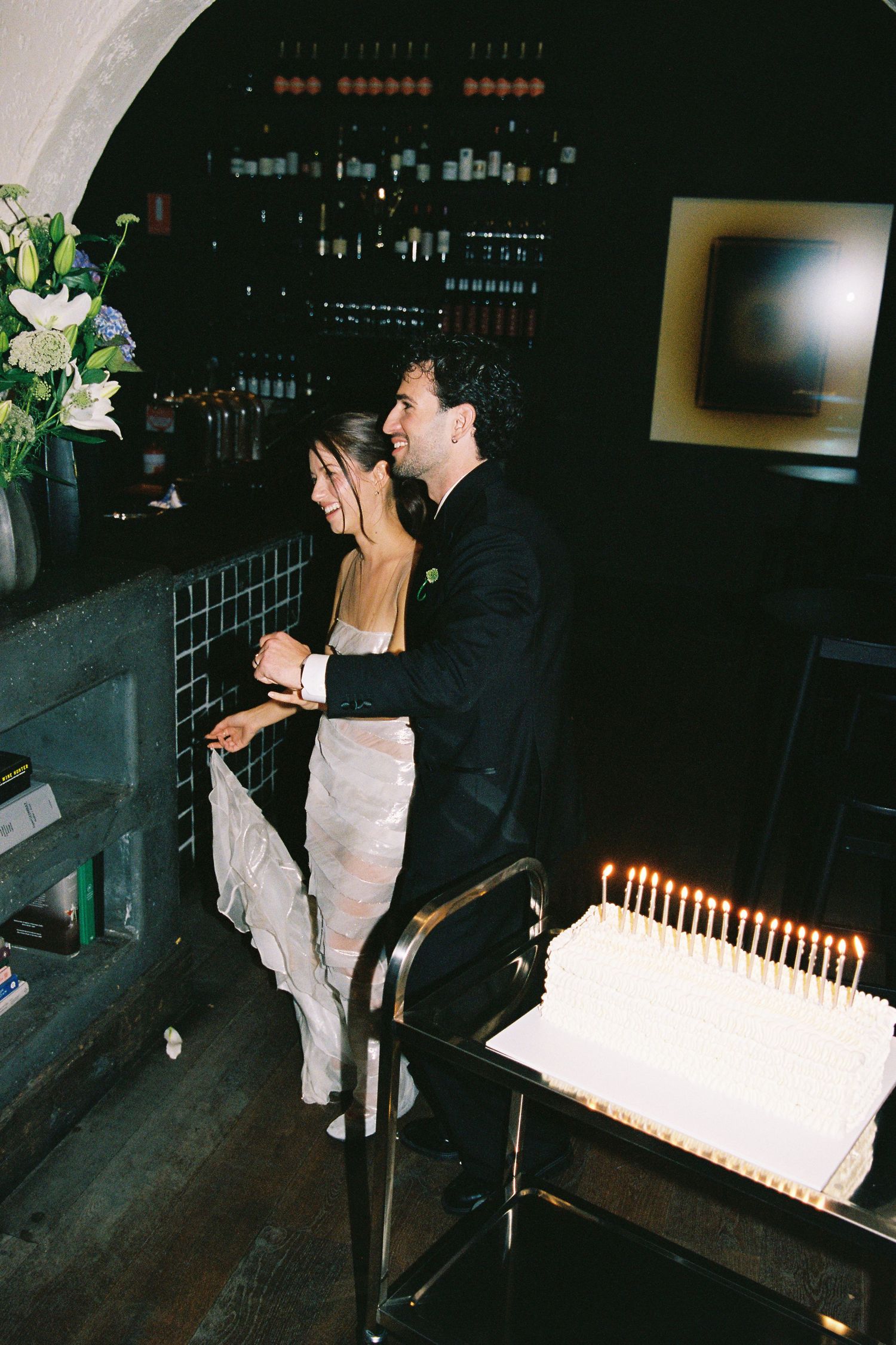Bride and groom cut white candlelit wedding cake together at dark intimate restaurant venue.