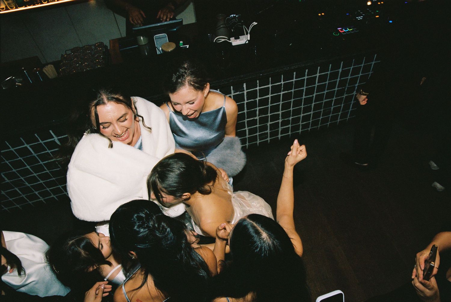 A group of women in formal attire gather and laugh together at a nighttime wedding event.