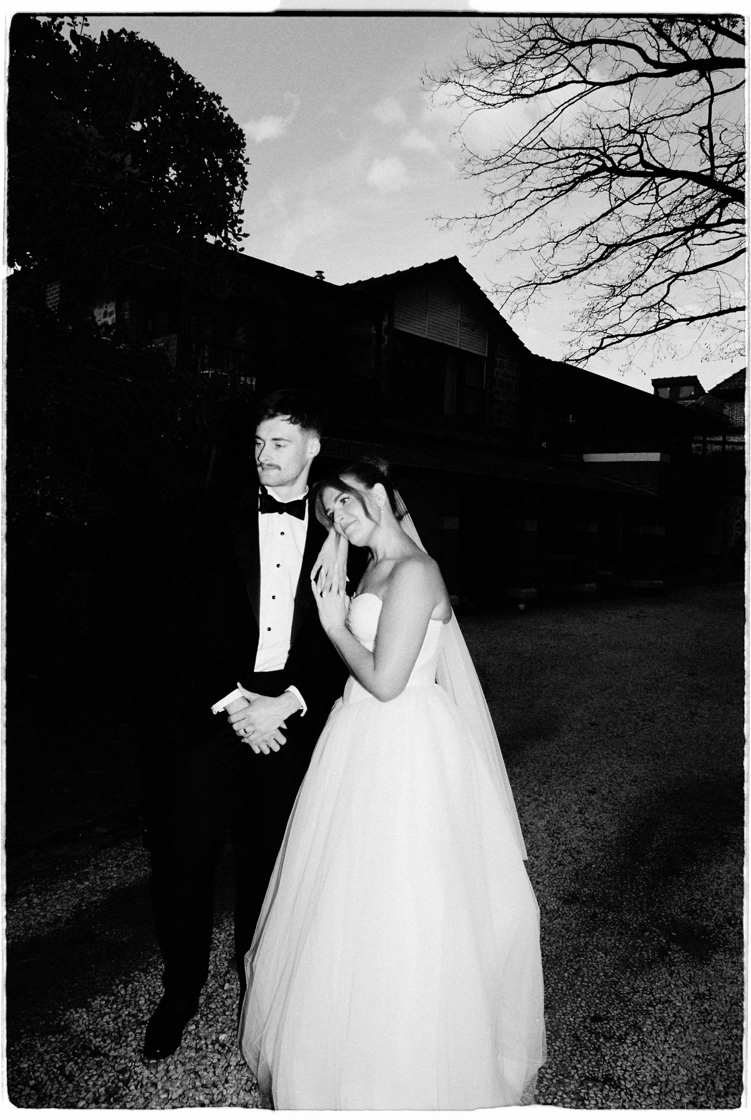 Black and white wedding photo of bride and groom standing outdoors at dusk near a rustic building.