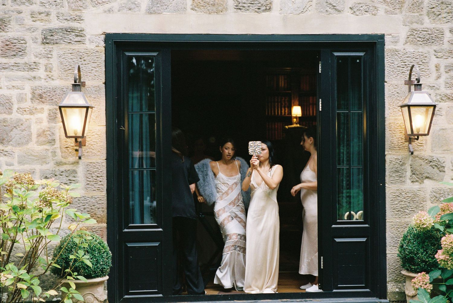 Bridesmaids in elegant white gowns exit through black double doors of stone wedding venue.