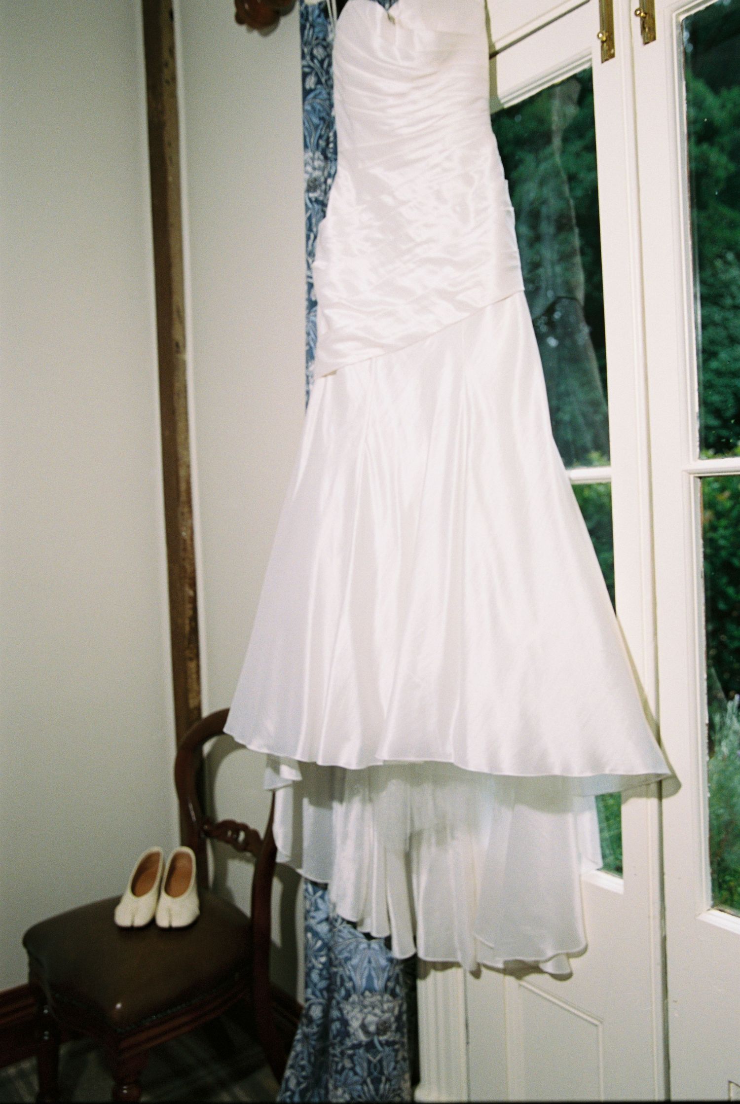 Elegant white mermaid wedding dress hangs by window with white bridal shoes on vintage wooden chair.