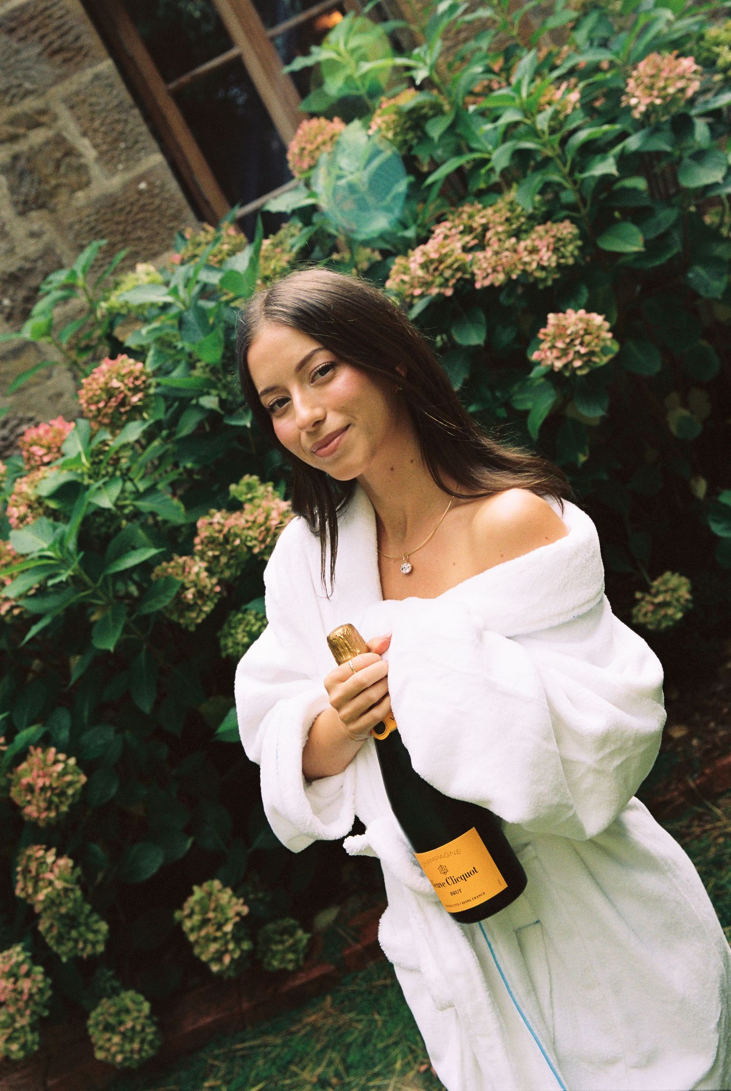 Bride in white robe holding Veuve Clicquot champagne bottle among blooming hydrangeas outdoors.